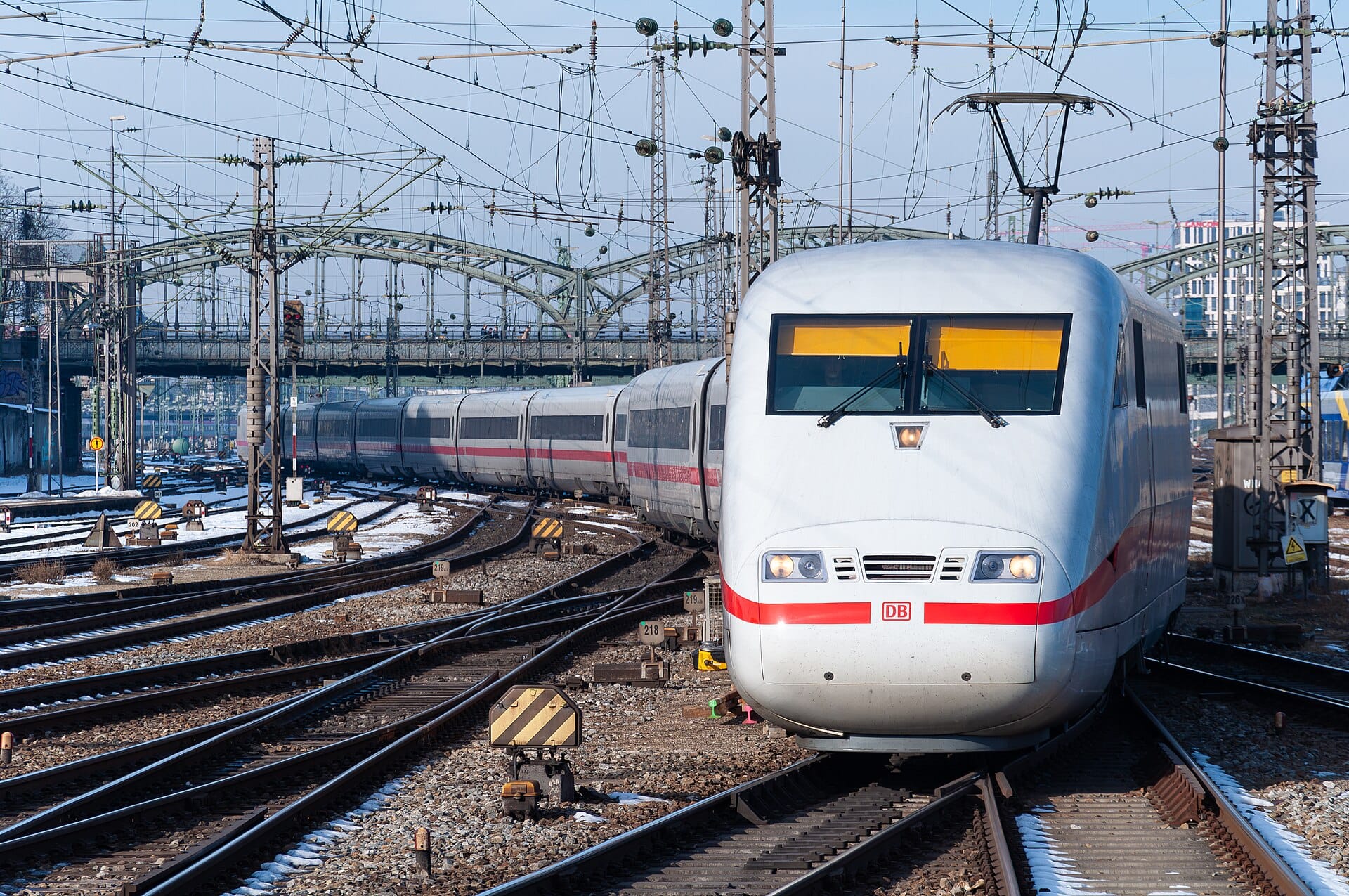 รถไฟ ICE สีขาวเข้าสู่ Munich Hauptbahnhof ที่ปกคลุมด้วยหิมะ ล้อมรอบด้วยรางและสายไฟเหนือหัวในวันฤดูหนาวที่แจ่มใส