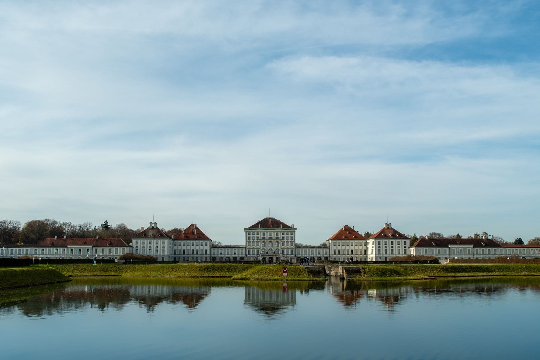 วิวกว้างของพระราชวัง Nymphenburg ในมิวนิก พร้อมน้ำสงบที่สะท้อนภาพพระราชวังประวัติศาสตร์และท้องฟ้ามีเมฆ
