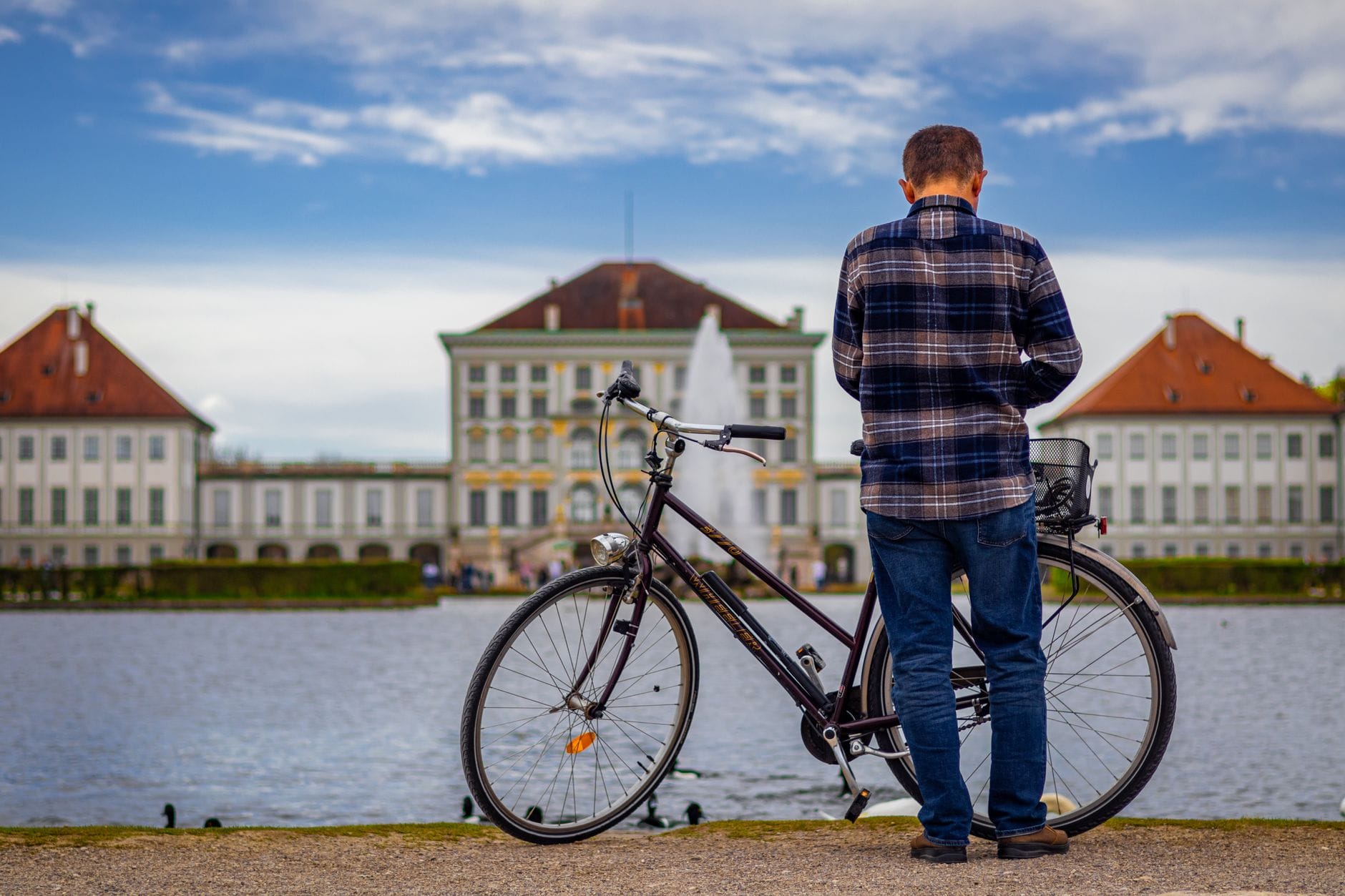 ชายคนหนึ่งพร้อมจักรยานยืนอยู่ใกล้แหล่งน้ำ มองไปยังพระราชวัง Nymphenburg ในเมือง Munich ใต้ท้องฟ้าสีฟ้าและเมฆ