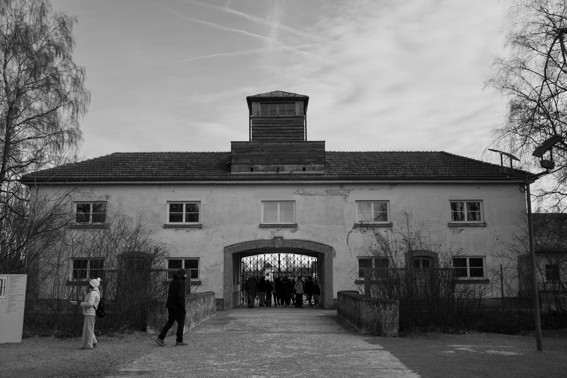ภาพถ่ายขาวดำของอาคารประตูทางเข้าของสถานที่รำลึก Dachau พร้อมนักท่องเที่ยวที่กำลังเดินเข้าไปใต้หอคอยยาม