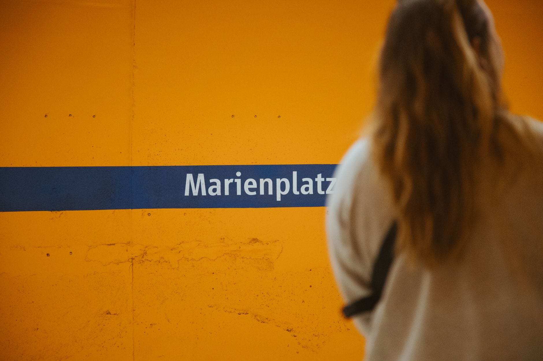 บุคคลยืนอยู่ที่สถานี S-Bahn Marienplatz ในเมืองมิวนิก พร้อมกำแพงสีส้มและชื่อสถานีสีน้ำเงินที่มองเห็นได้ชัดเจน
