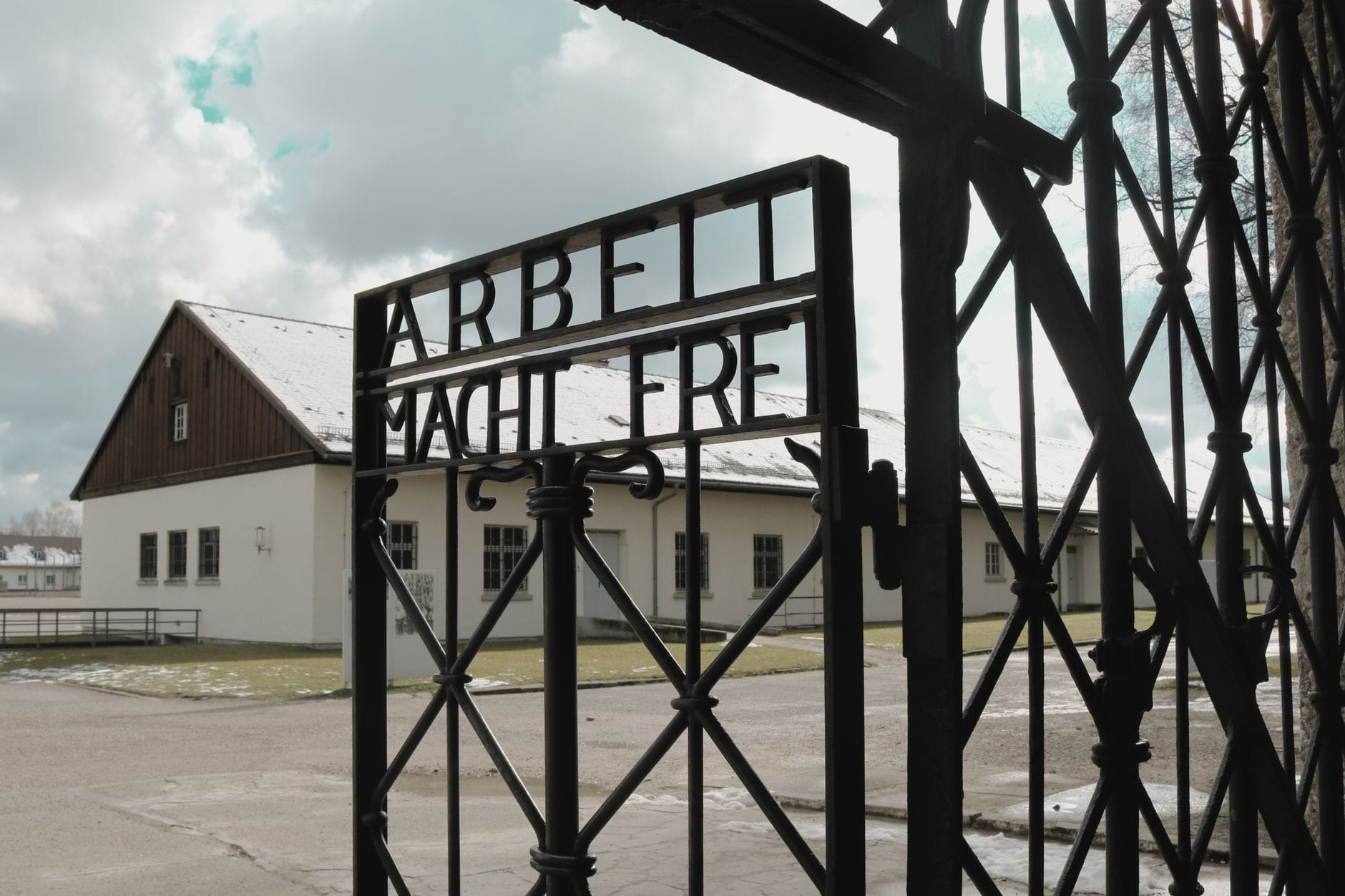 วิวผ่านประตูเหล็กดัดที่มีจารึก 'Arbeit macht frei' ที่ค่ายกักกัน Dachau โดยมีค่ายนักโทษอยู่เบื้องหลังในวันที่มีเมฆมาก