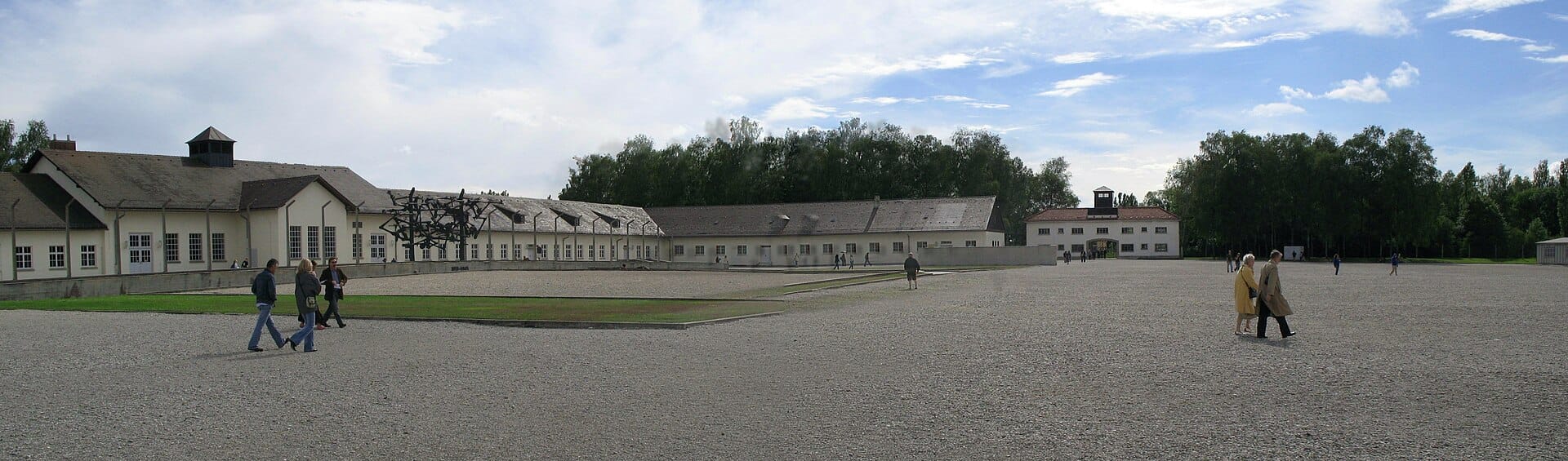 มุมมองกว้างของสถานที่อนุสรณ์ค่ายกักกัน Dachau พร้อมลานสวนสนามขนาดใหญ่ อาคารบำรุงรักษา และประตูทางเข้า ใต้ท้องฟ้ามีเมฆบางส่วน
