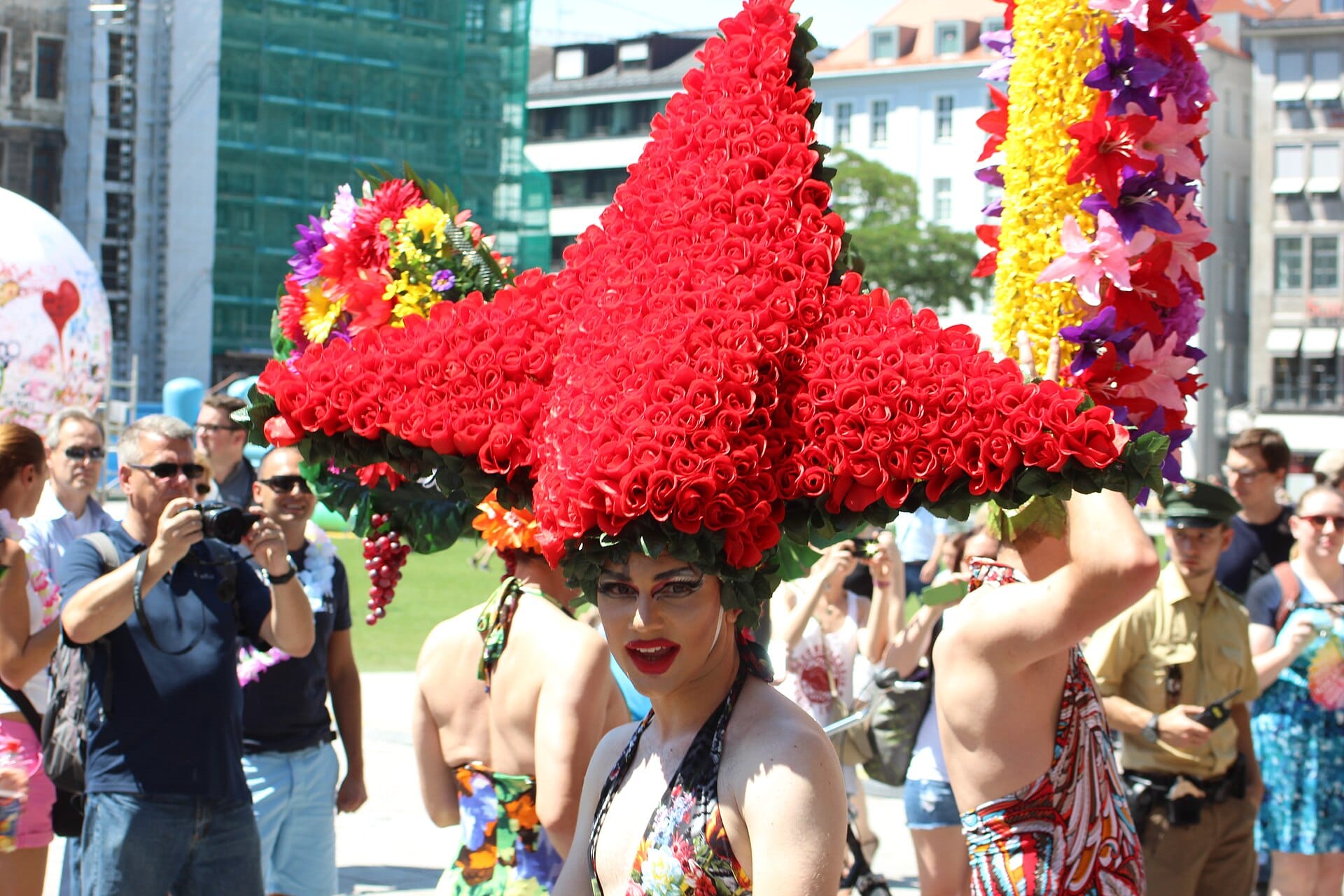 นักแสดงดรากสวมหมวกดอกไม้สีแดงประดับอย่างวิจิตรในงาน Pride Parade กลางแจ้งใจกลางเมือง München โดยมีผู้ชมและอาคารของเมืองเป็นฉากหลัง