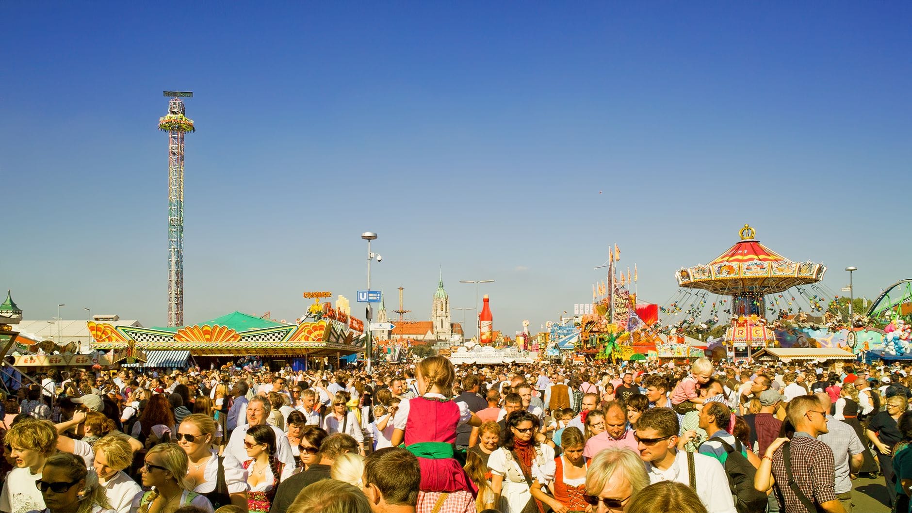 ฝูงชนจำนวนมากในงาน Oktoberfest พร้อมเครื่องเล่นสวนสนุก ร้านอาหาร และผู้คนในชุดพื้นเมืองบาวาเรียในวันที่แดดจัด