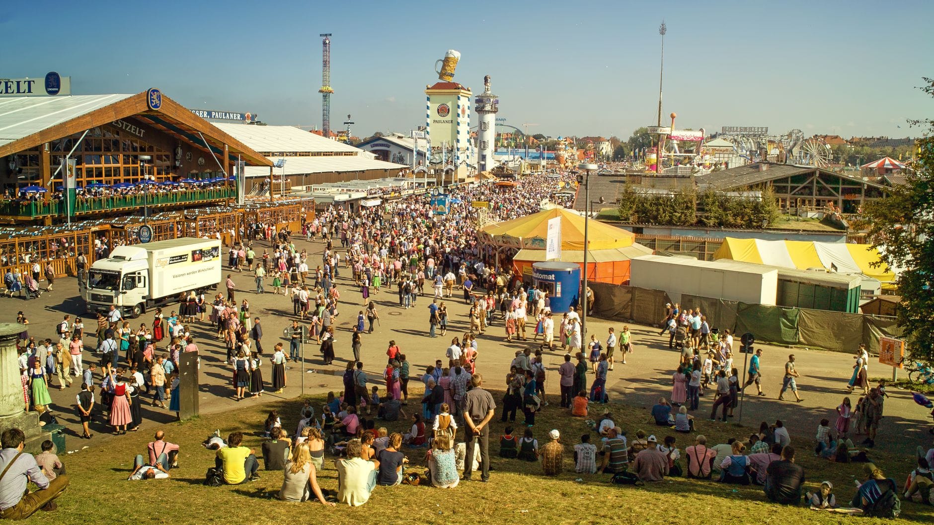 ฝูงชนงาน Oktoberfest เมือง München และเต็นท์เบียร์ที่สนาม Theresienwiese ในวันที่มีแดดจัด พร้อมผู้คนที่นั่งและเดินเล่นรอบๆ สถานที่ท่องเที่ยวของเทศกาล