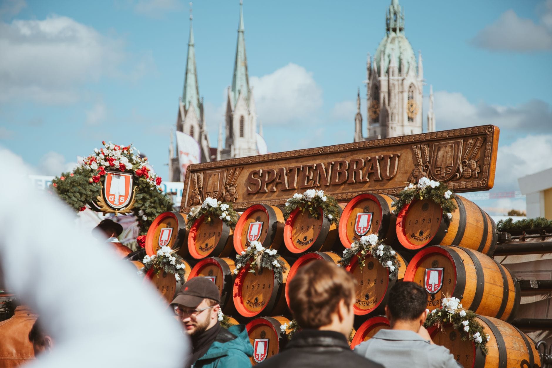 ผู้คนรวมตัวกันรอบกองถังเบียร์ Spatenbräu ในงาน Oktoberfest โดยมียอดแหลมของโบสถ์เมือง München เป็นฉากหลัง