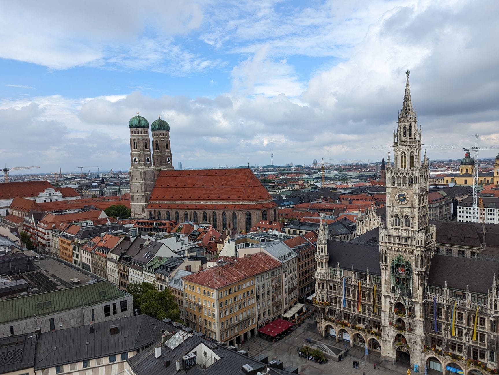 ภาพมุมกว้างใจกลางเมืองมิวนิก เห็น Neues Rathaus, Frauenkirche และอาคารหลังคาแดงใต้ท้องฟ้ามีเมฆ