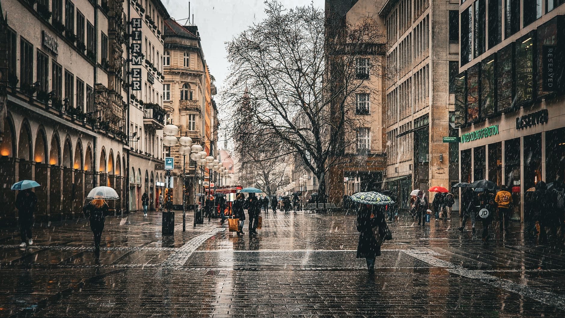 ถนนช้อปปิ้งสำหรับคนเดินเท้าที่คึกคักใน München โดยมีผู้คนถือร่มในวันที่ฝนตก รายล้อมด้วยร้านค้าและอาคารประวัติศาสตร์