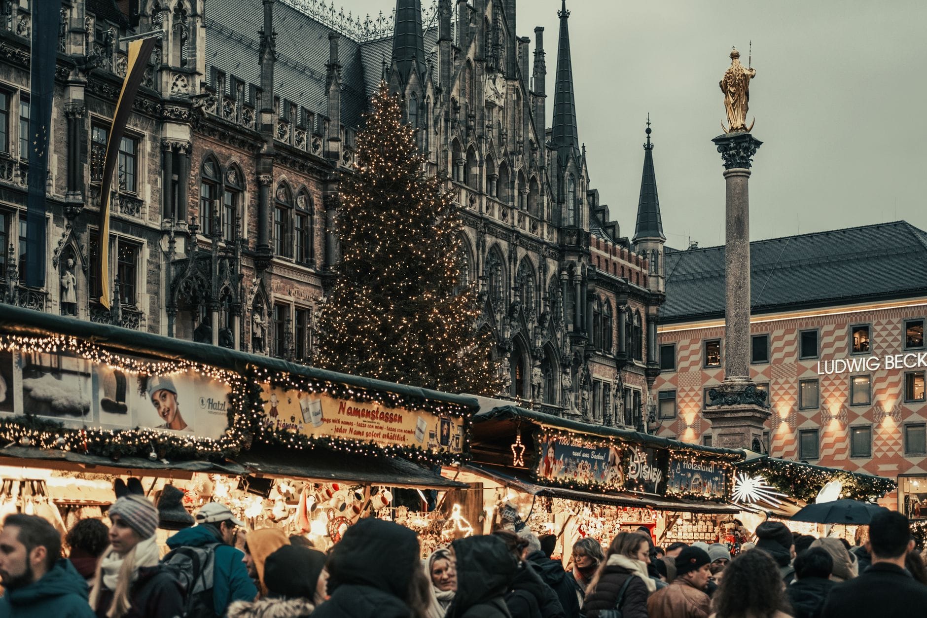 ผู้คนเดินเลือกซื้อของที่แผงตลาดบริเวณ Marienplatz ในฤดูหนาว โดยมีศาลาว่าการใหม่และเสา Mariensäule ทองคำเป็นฉากหลัง