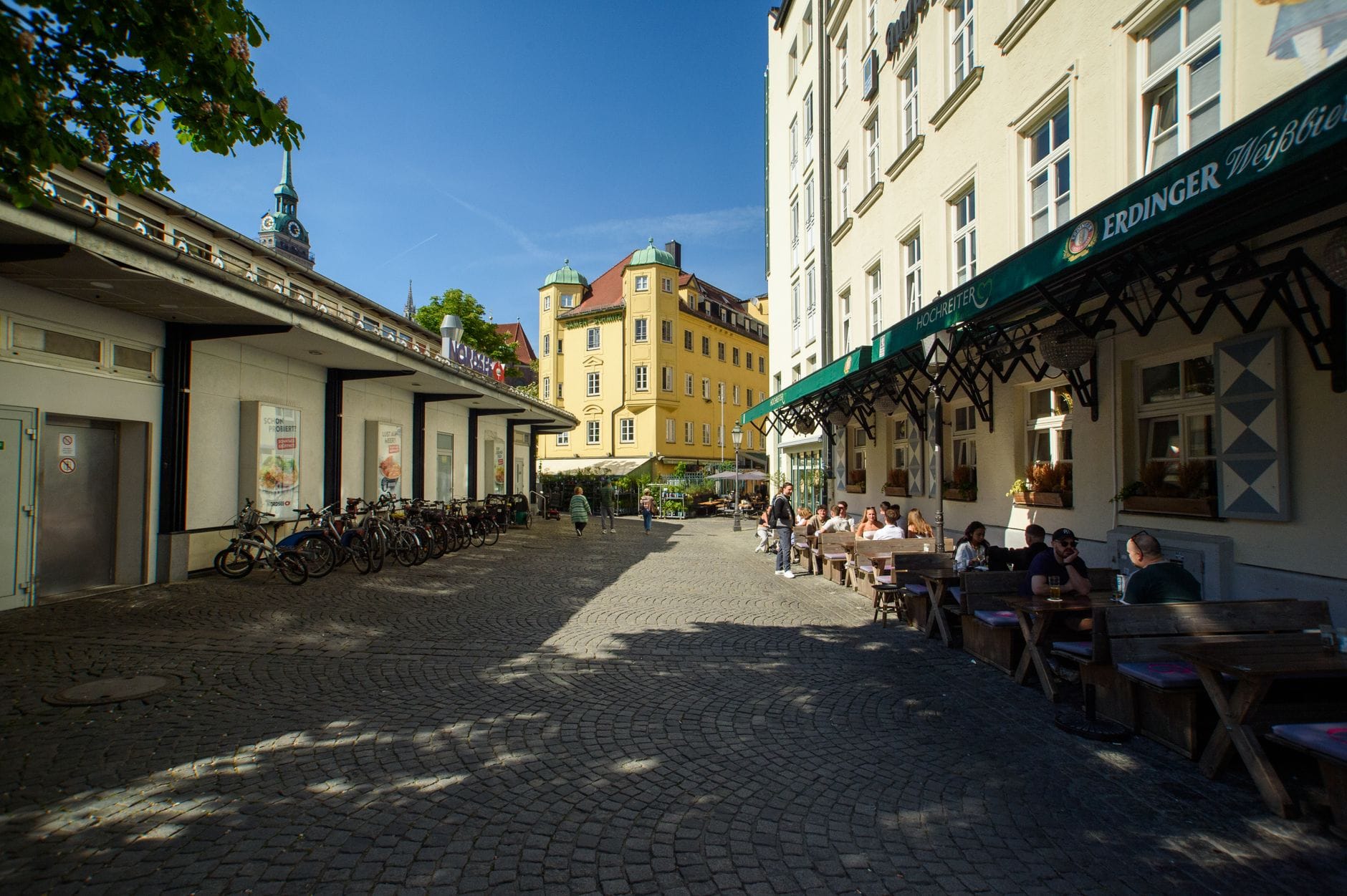 ภาพถนนในย่าน Gärtnerplatz เมือง Munich ในวันที่แดดสดใส มีผู้คนนั่งที่โต๊ะกลางแจ้งและร้านค้าในอาคารประวัติศาสตร์สีสันสวยงาม