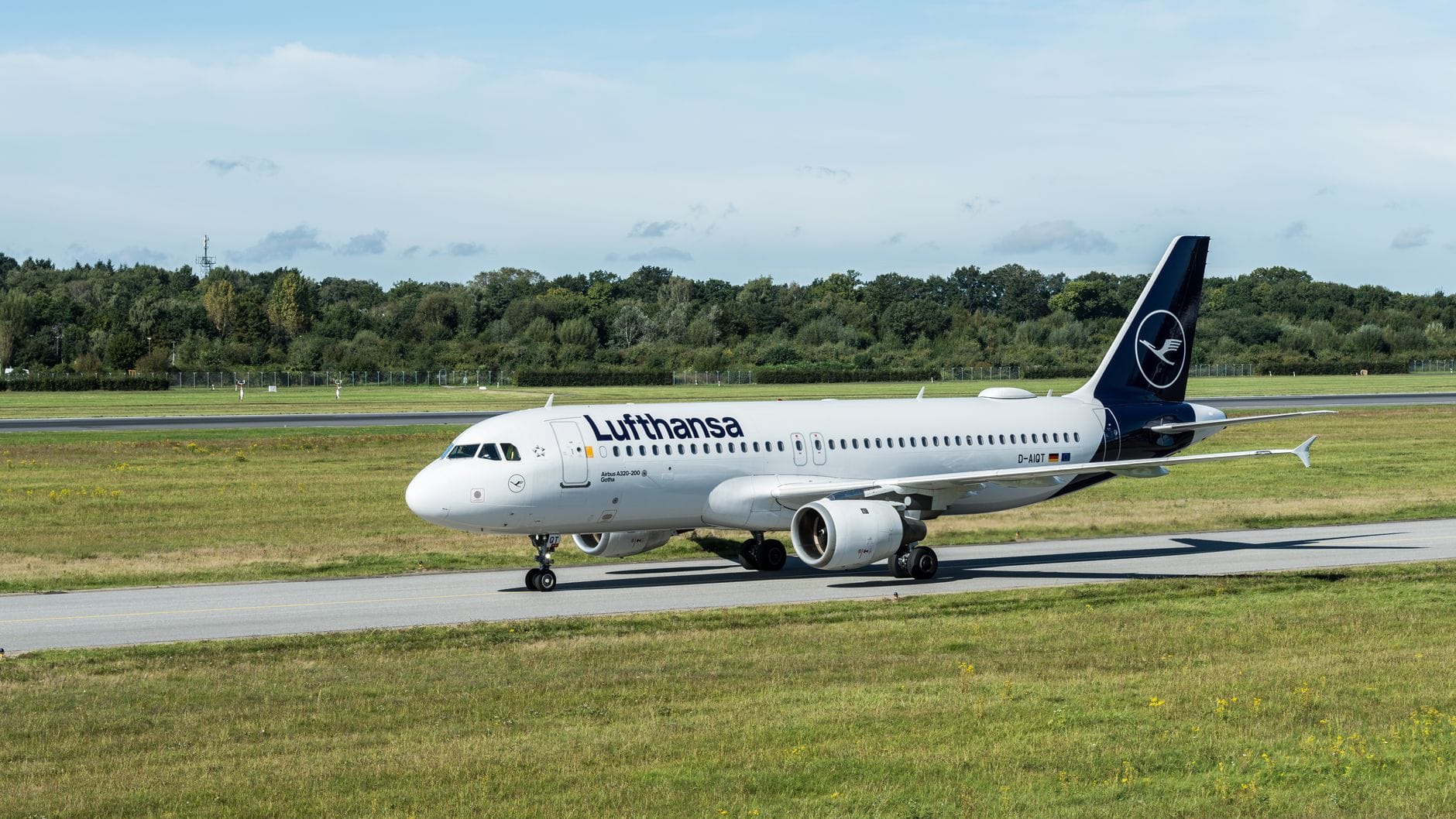 เครื่องบิน Lufthansa กำลังแล่นบนรันเวย์ที่มีหญ้าสีเขียว ท้องฟ้าสีฟ้า และต้นไม้อยู่เบื้องหลัง