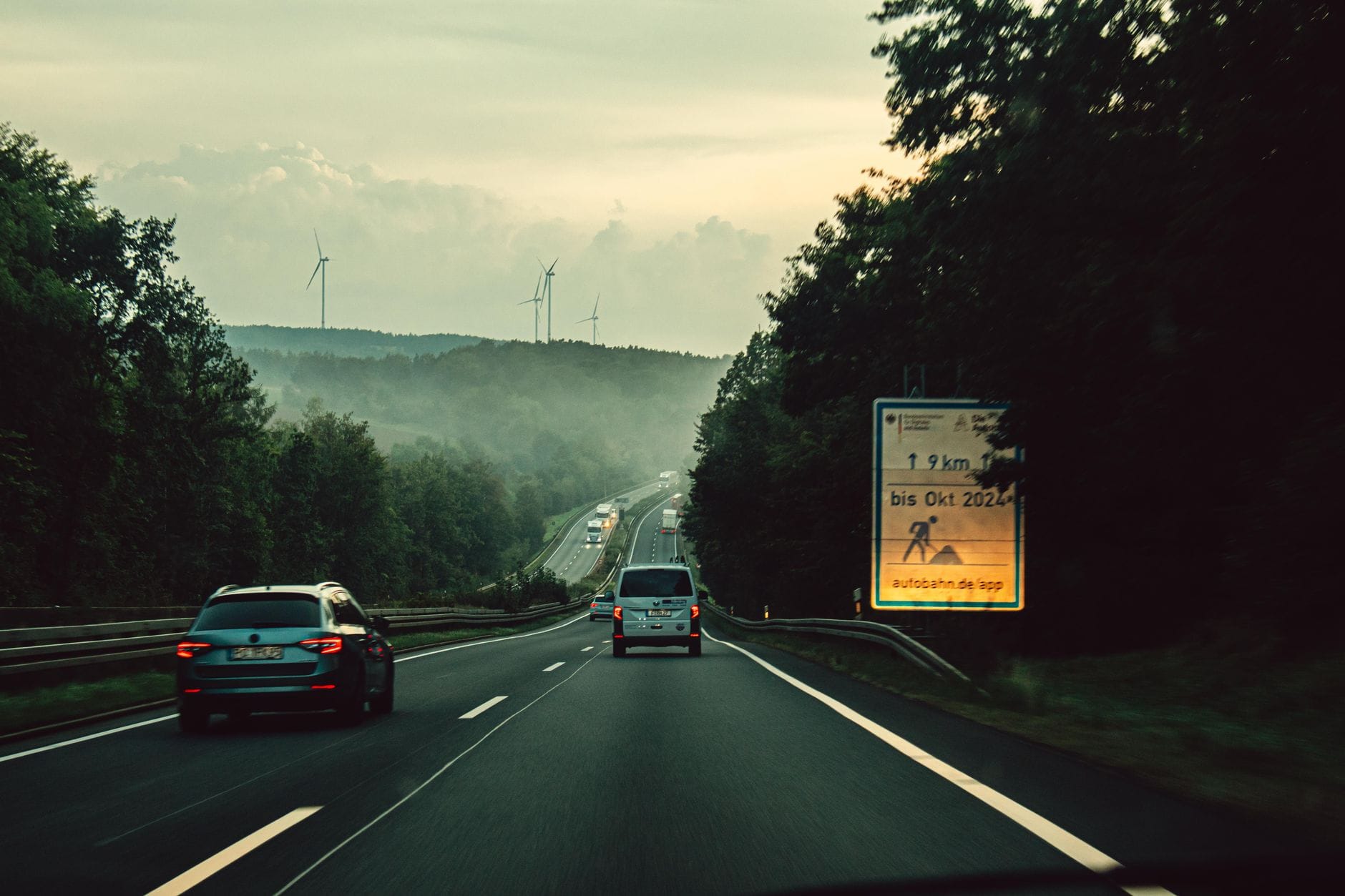 รถยนต์วิ่งบน Autobahn ของเยอรมนีผ่านเนินเขาที่เต็มไปด้วยป่าไม้ มองจากมุมมองของผู้ขับขี่โดยมีกังหันลมอยู่เบื้องหลัง