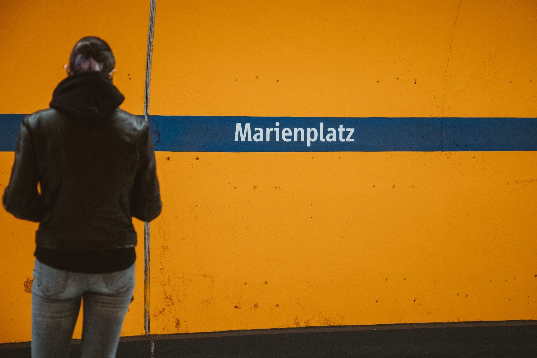 บุคคลยืนอยู่หน้ากำแพงสีส้มสดใสที่สถานี U-Bahn Marienplatz ในมิวนิก พร้อมป้ายชื่อสถานี