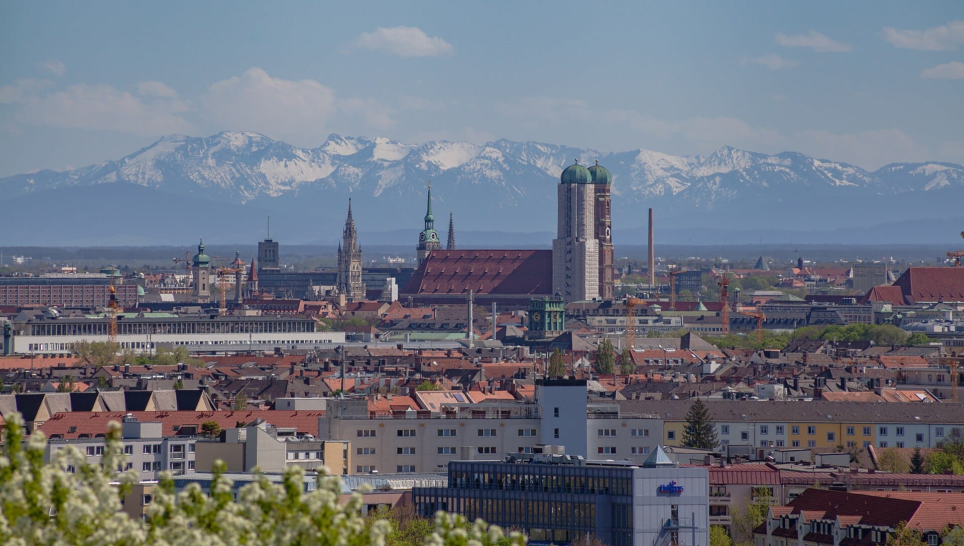 ภาพพาโนรามาของเมืองมิวนิกพร้อม Frauenkirche และเมืองเก่า มีเทือกเขา Alps ปกคลุมด้วยหิมะเป็นฉากหลังใต้ท้องฟ้าสีฟ้า