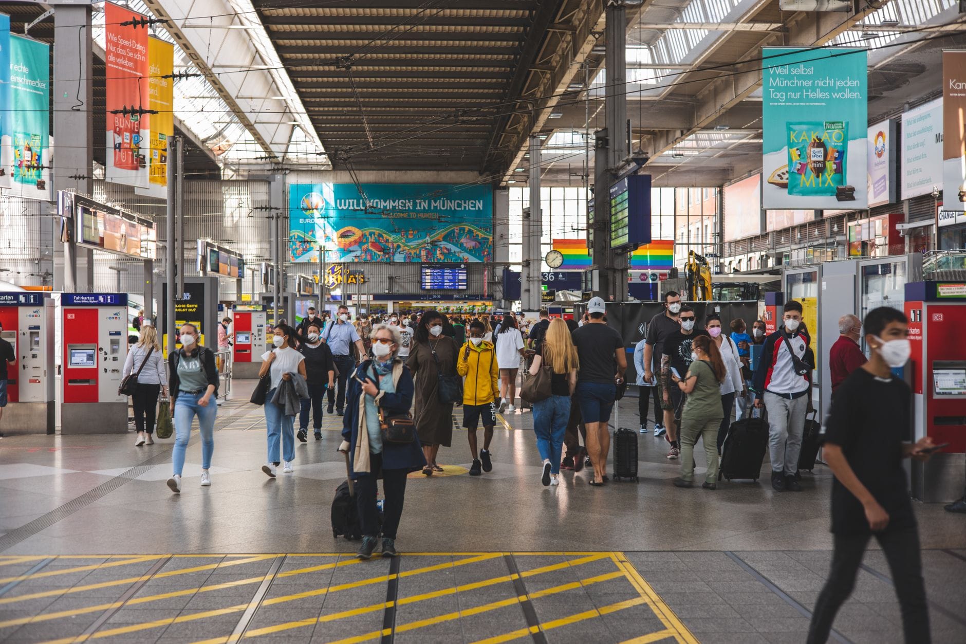 ภายในสถานีรถไฟสมัยใหม่ที่คึกคักพร้อมนักเดินทางและป้ายต้อนรับขนาดใหญ่ที่เขียนว่า 'Willkommen in München'