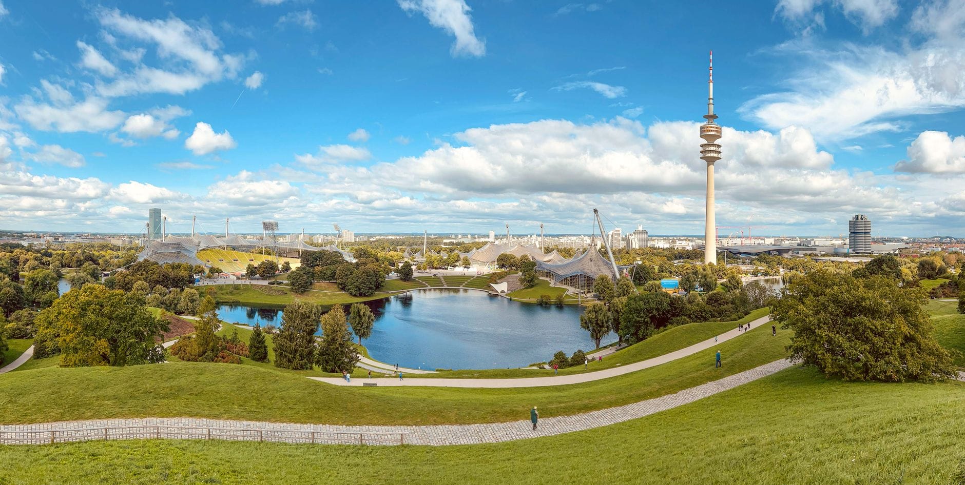 วิวพาโนรามาของ Olympiapark ในมิวนิก เห็นทะเลสาบ ทางเดินกว้าง หลังคาโมเดิร์นของสนามกีฬาโอลิมปิก และหอคอยโอลิมปิกโดดเด่นบนท้องฟ้าสีฟ้ากับเมฆขาวฟุ้ง