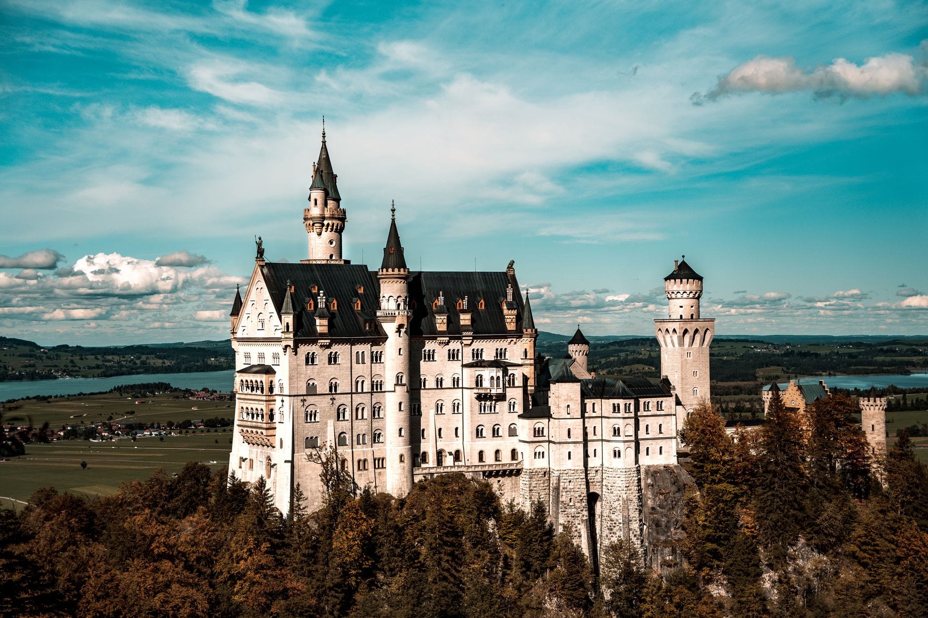 ปราสาท Neuschwanstein อันงดงามตั้งอยู่บนเนินเขาที่ปกคลุมด้วยป่าไม้ โดยมีทุ่งนาสีเขียวและทะเลสาบเป็นฉากหลังใต้ท้องฟ้าสีฟ้า