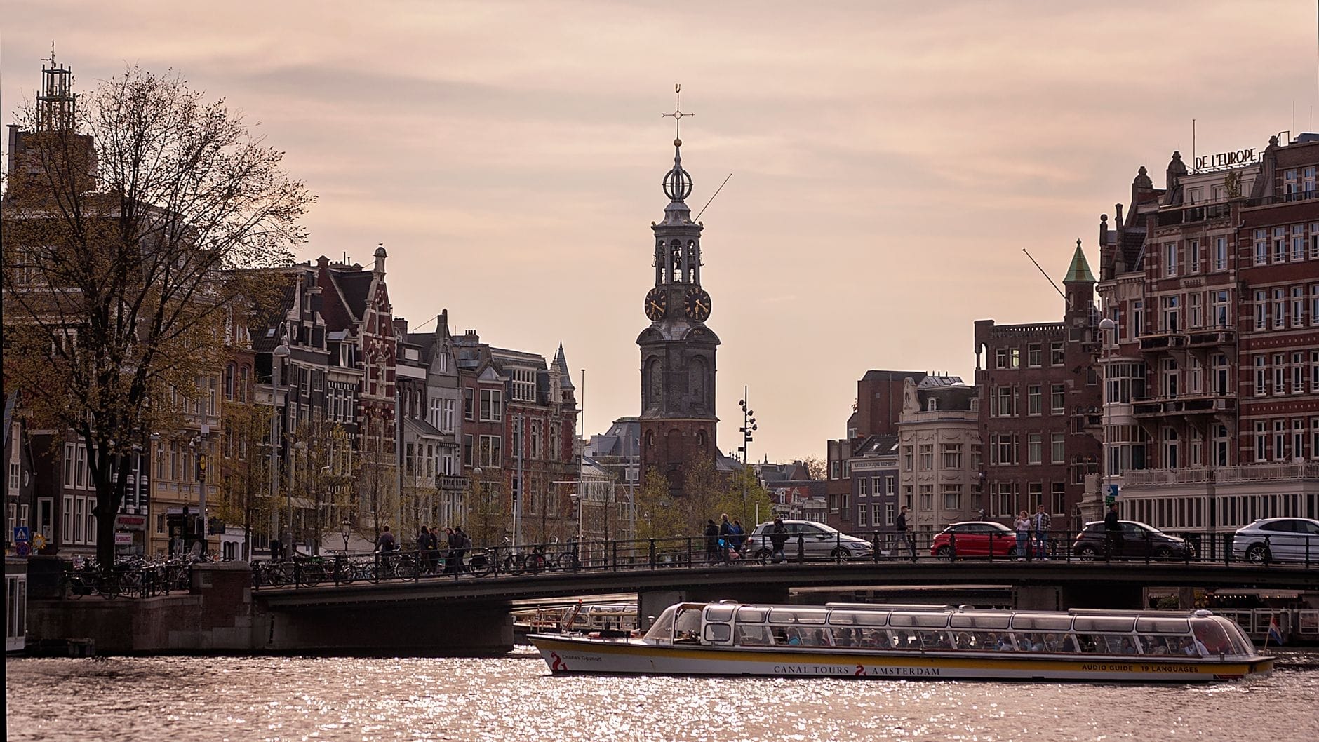 Vista de la Munttoren (Torre de la Moneda) en Ámsterdam, elevándose sobre las casas del canal y un animado puente con un barco turístico en el río Amstel al atardecer.