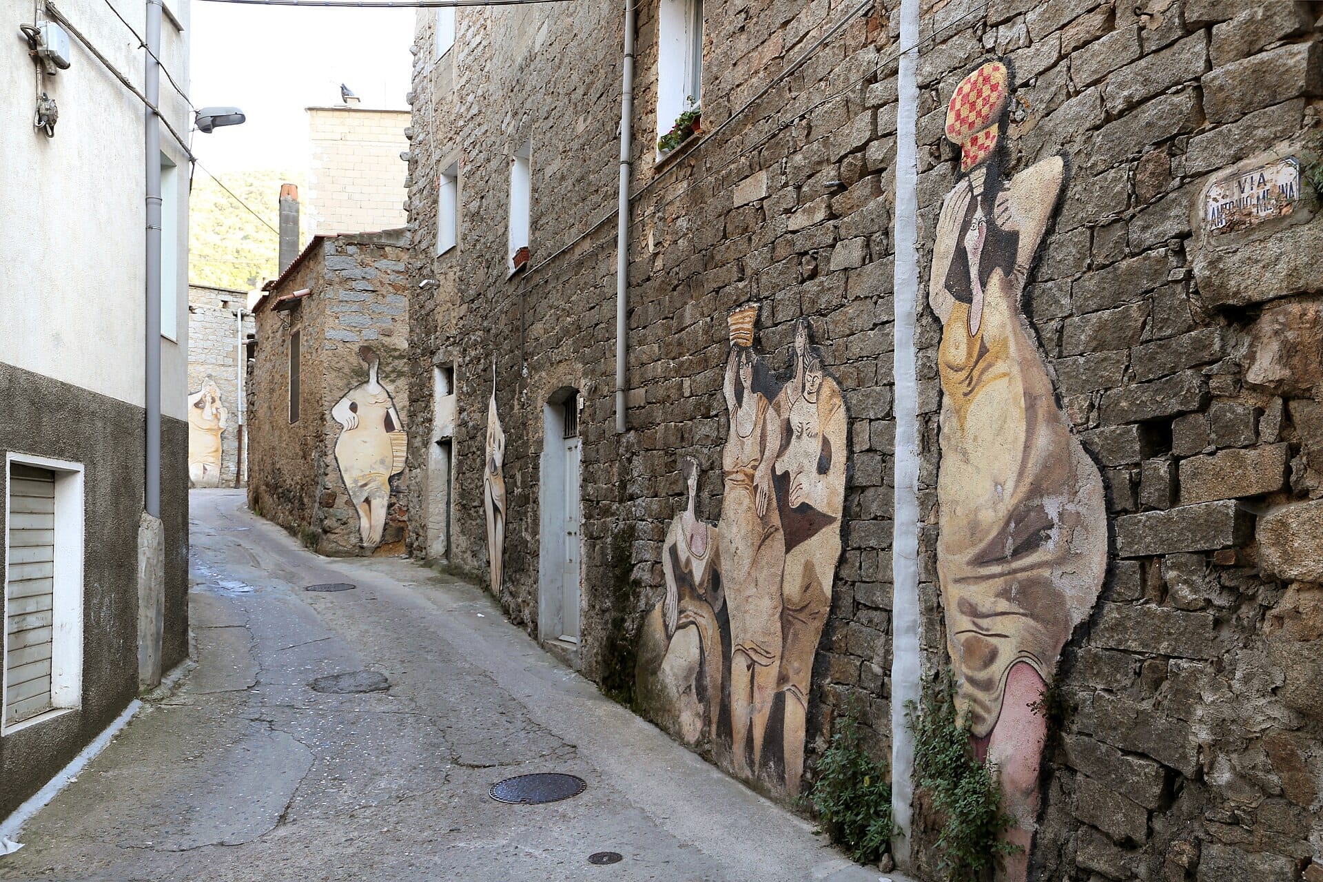 Straßenansicht der Steingebäude von Orgosolo, die mit großen, stilisierten Wandmalereien von Menschen verziert sind, Teil der berühmten Murales di Orgosolo.