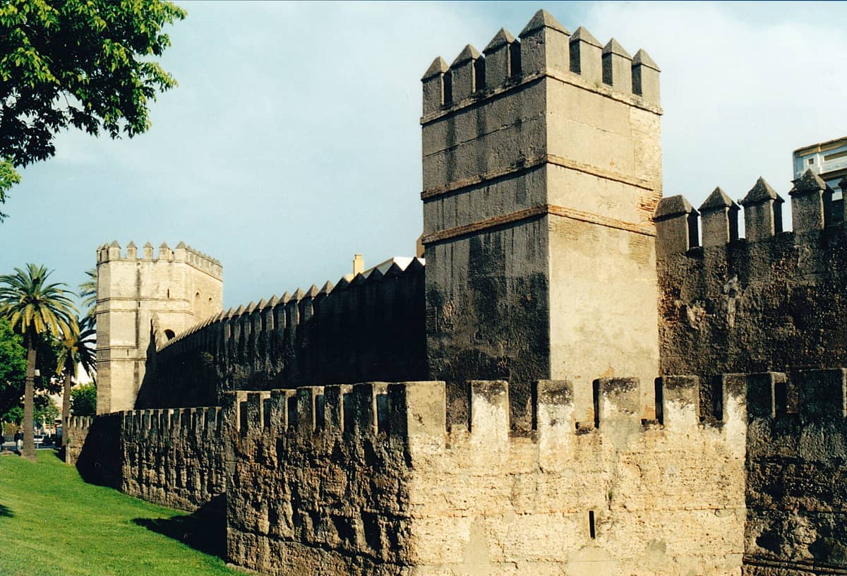 หอคอยหินและกำแพงยุคกลางที่มีใบปราการของ Murallas de Sevilla ภายใต้ท้องฟ้าแจ่มใสพร้อมต้นปาล์มอยู่ใกล้เคียง