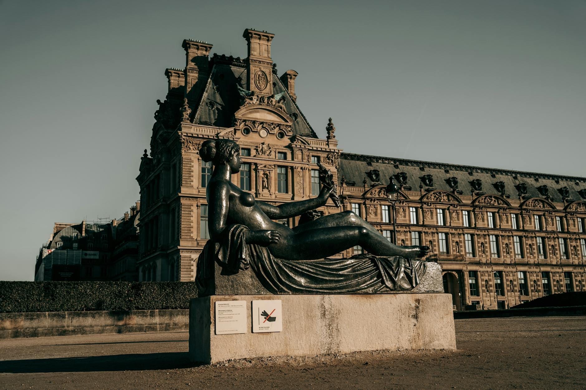 Grande scultura bronzea di figura femminile distesa, collocata all'aperto davanti a un elegante edificio parigino storico sotto un cielo limpido, che evoca l'atmosfera di un museo a cielo aperto.