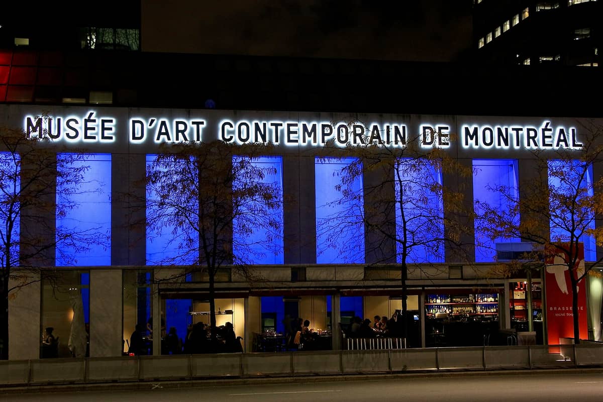 Nachtansicht der Fassade des Musée d'art contemporain de Montréal mit beleuchteten blauen Fenstern und dem leuchtenden Museumsnamen über dem Eingang.