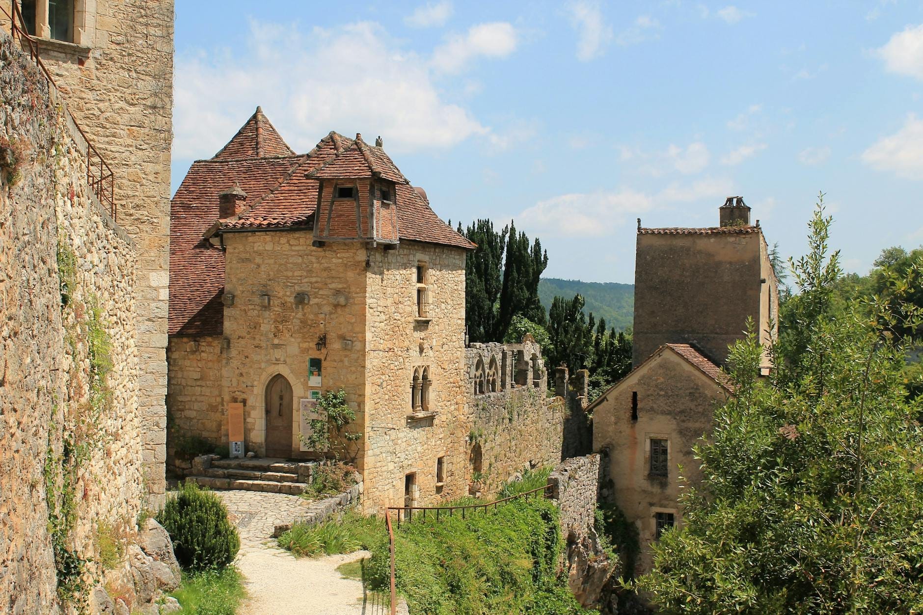 Pittoreschi edifici medievali in pietra con tetti spioventi e un viottolo stretto, immersi in un paesaggio verdeggiante su un colle soleggiato.
