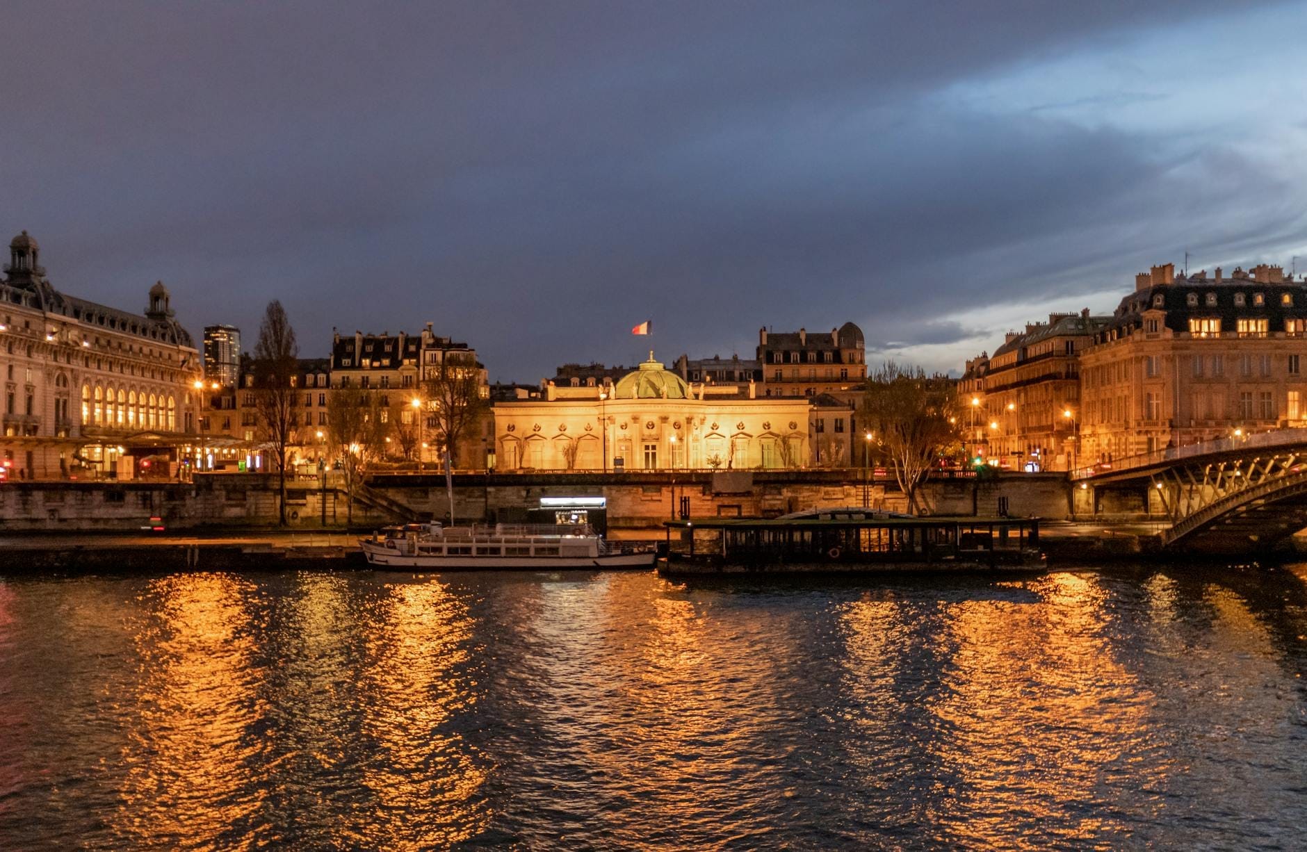 L'elegante edificio del museo illuminato al crepuscolo, visto dalla Senna, con i riflessi luminosi sull'acqua e lo skyline di Parigi sullo sfondo.