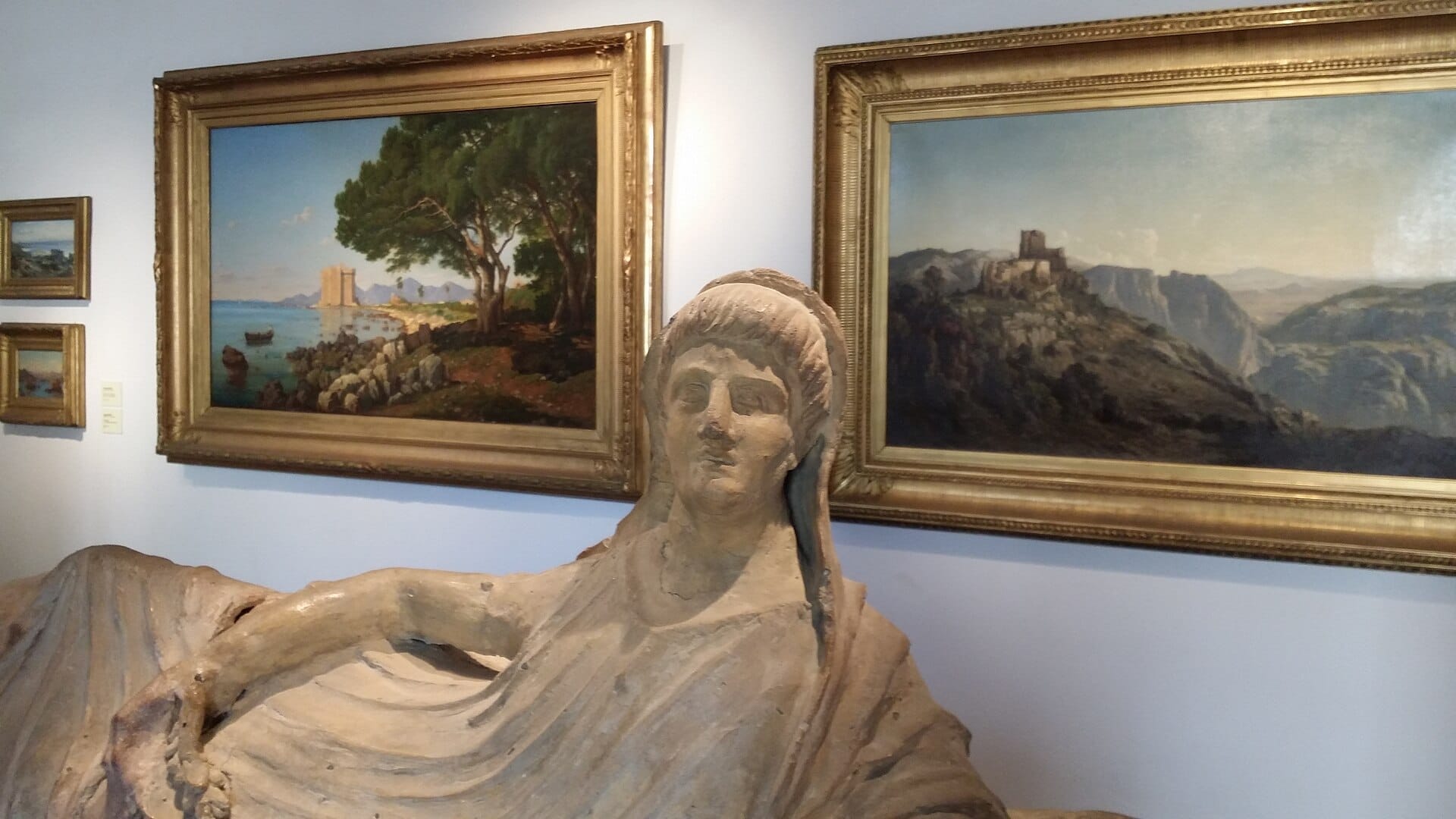 Statua in pietra di una figura sdraiata esposta davanti a dipinti di paesaggi incorniciati al Musée des Explorations du monde di Cannes.