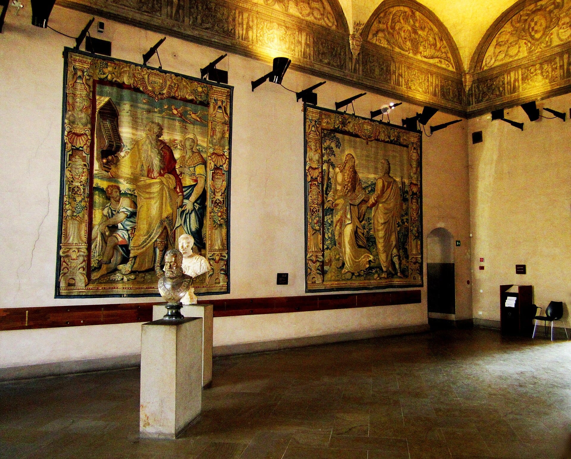 Galleria interna dei Musei del Castello Sforzesco con due arazzi rinascimentali ornati, busti in marmo, soffitto ad arco e illuminazione calda.