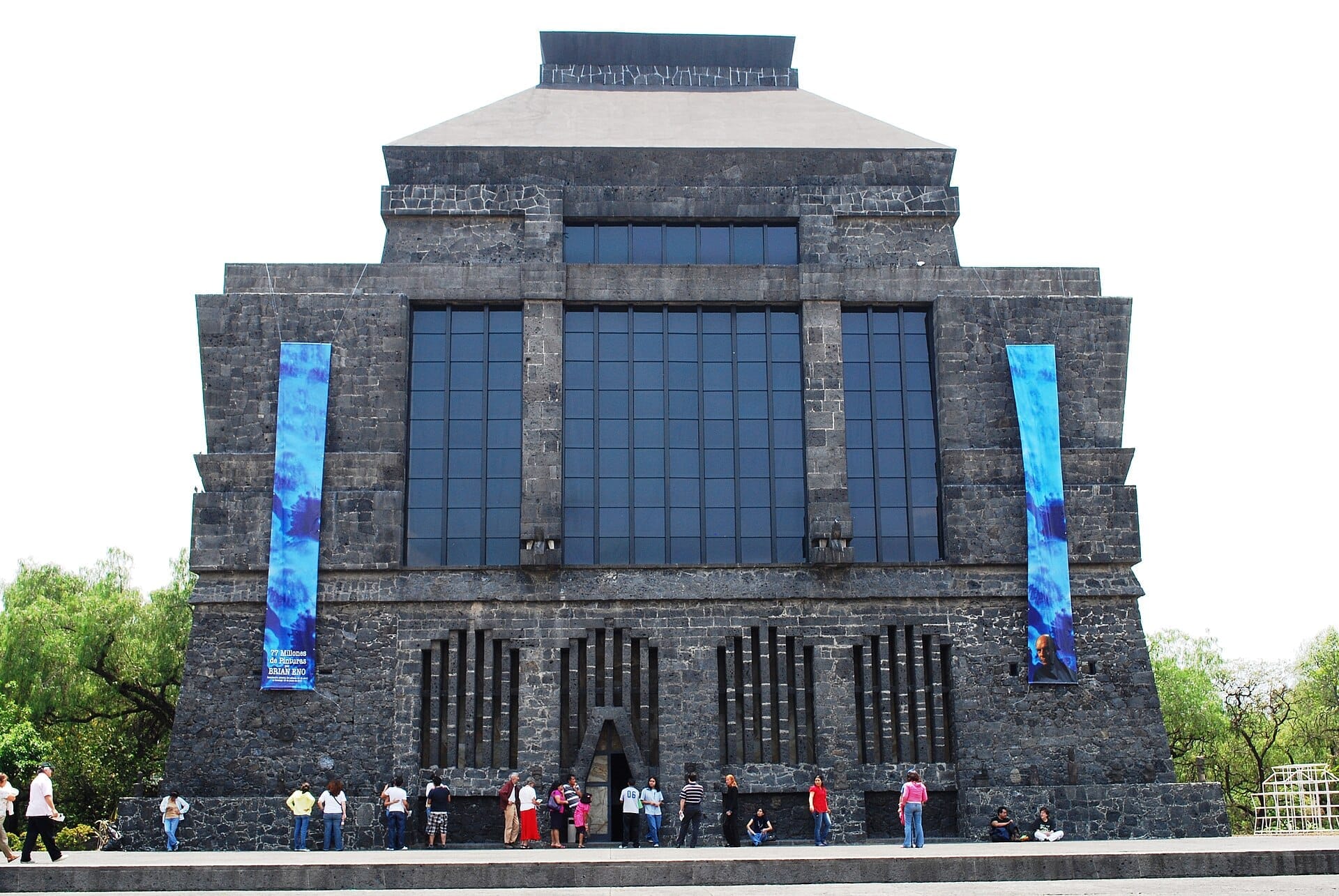 Vista frontale del Museo Anahuacalli che mostra la sua facciata in pietra vulcanica, grandi finestre, striscioni blu e visitatori in piedi fuori in una giornata di sole.