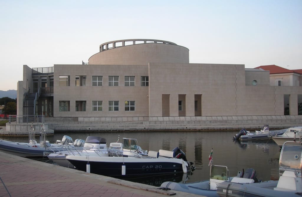 Frontansicht des Museo Archeologico di Olbia, ein modernes beigefarbenes Museumsgebäude mit geometrischen Linien, das sich im ruhigen Wasser des alten Hafens spiegelt.