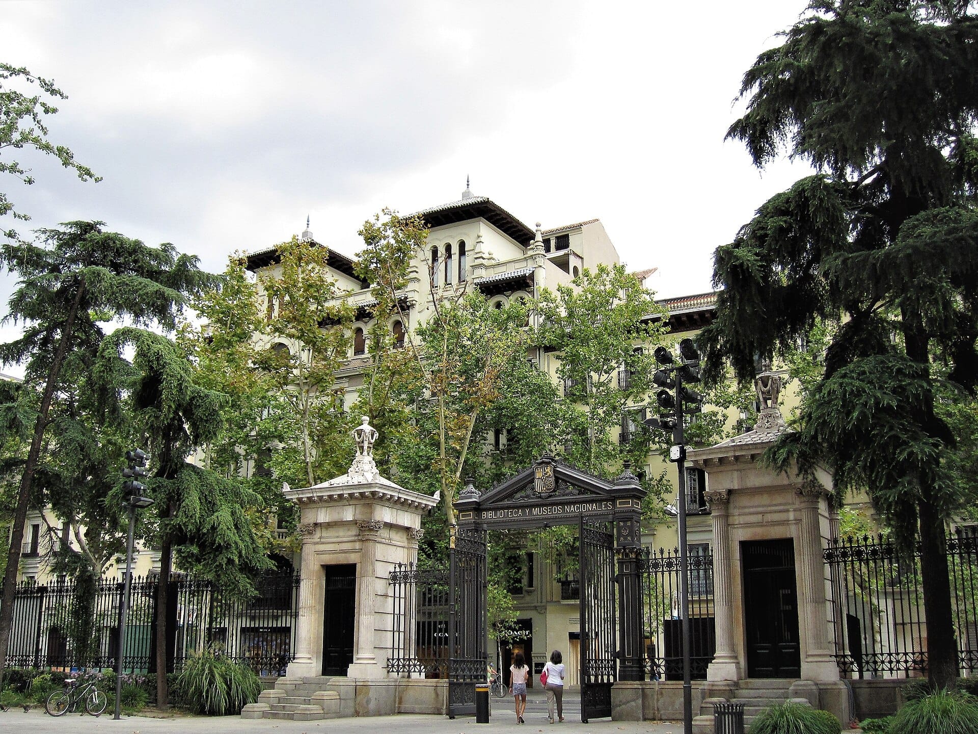 Il grande portale d'ingresso e la storica facciata del Museo Arqueológico Nacional a Madrid, incorniciati da rigogliosi alberi verdi in una giornata nuvolosa.