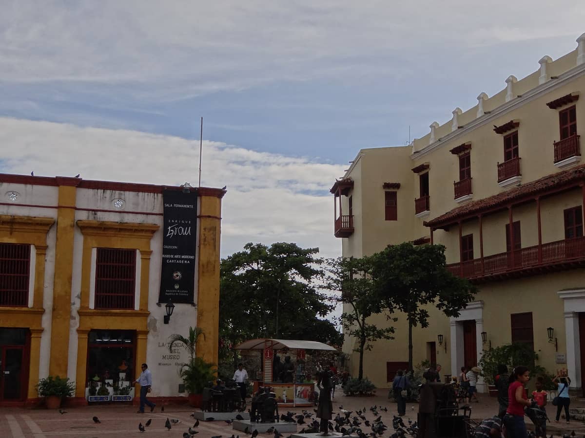 Colonial-era buildings housing the Museo de Arte Moderno de Cartagena with people, trees, and pigeons in Plaza de San Pedro Claver.