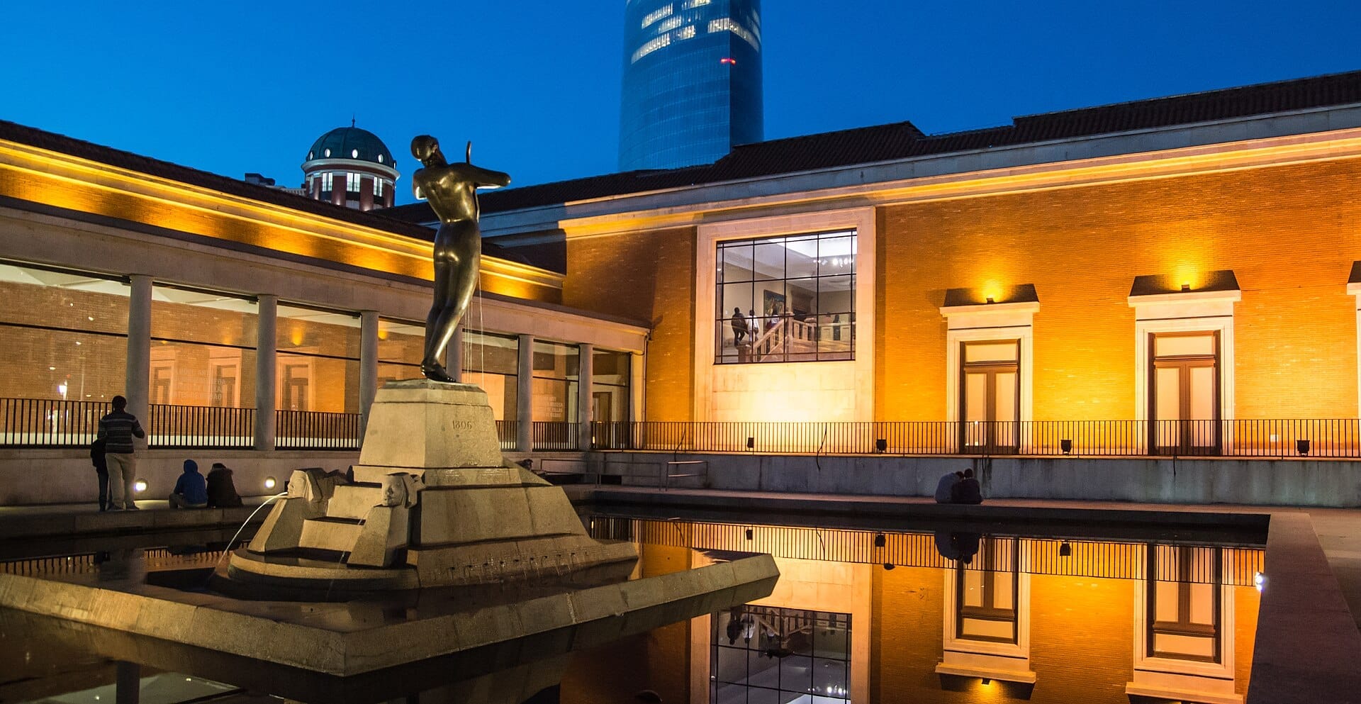 Evening view of Museo de Bellas Artes de Bilbao with its brick facade brightly lit, statue and reflecting pool in the foreground.
