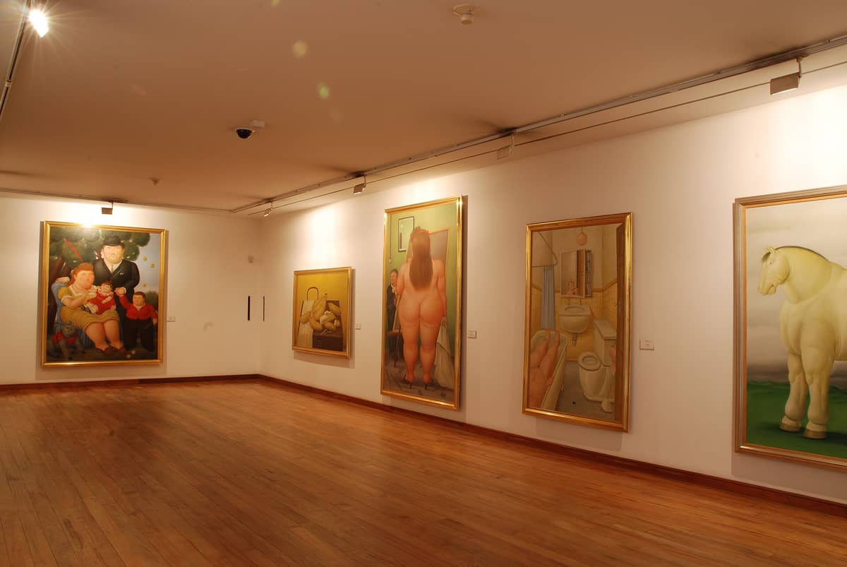 Interior view of Museo Botero showing several iconic Botero paintings on the walls, including voluminous figures, with warm lighting and wooden floors.
