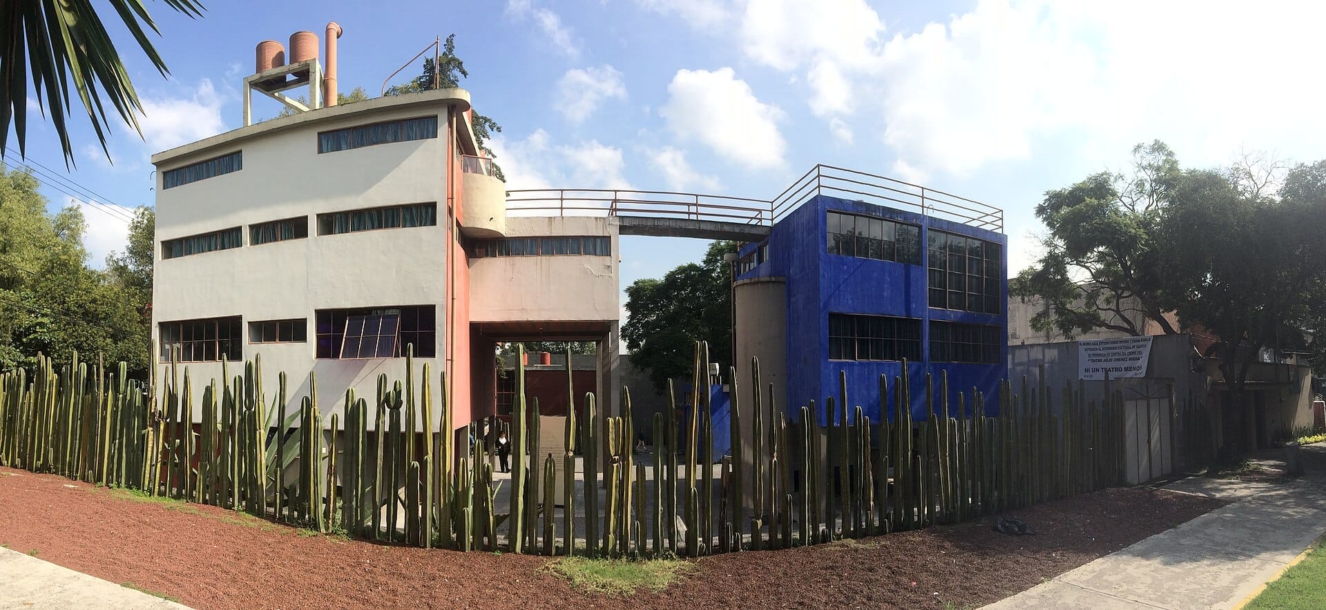 Museo Casa Estudio Diego Rivera y Frida Kahlo con i suoi edifici modernisti collegati in rosso, bianco e blu e un alto recinto di cactus sotto un cielo soleggiato.