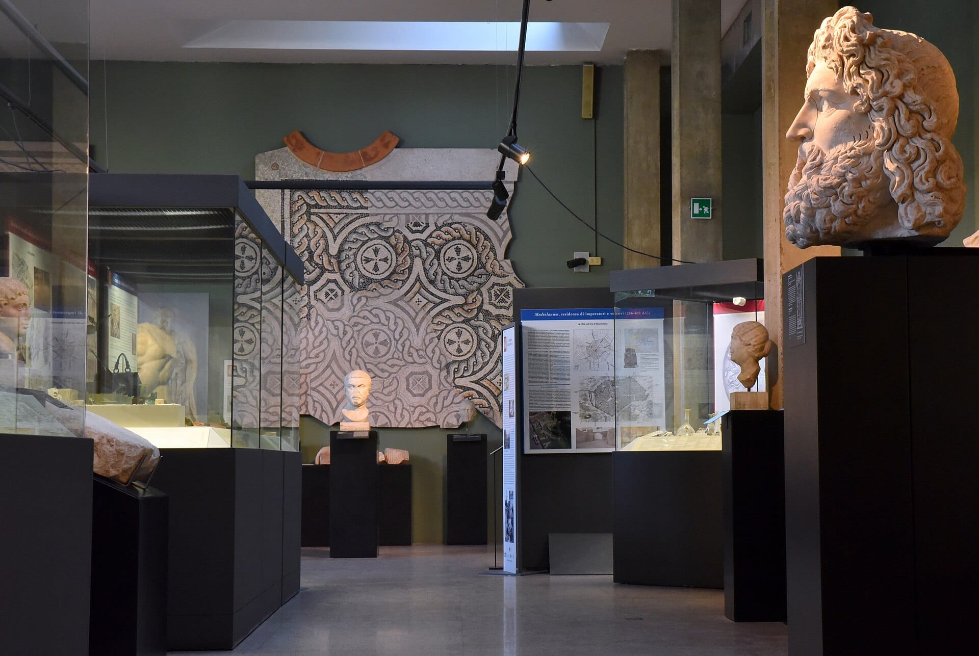 Sala espositiva del Museo Civico Archeologico di Milano con busti romani, pannelli informativi e un grande mosaico decorativo sulla parete.