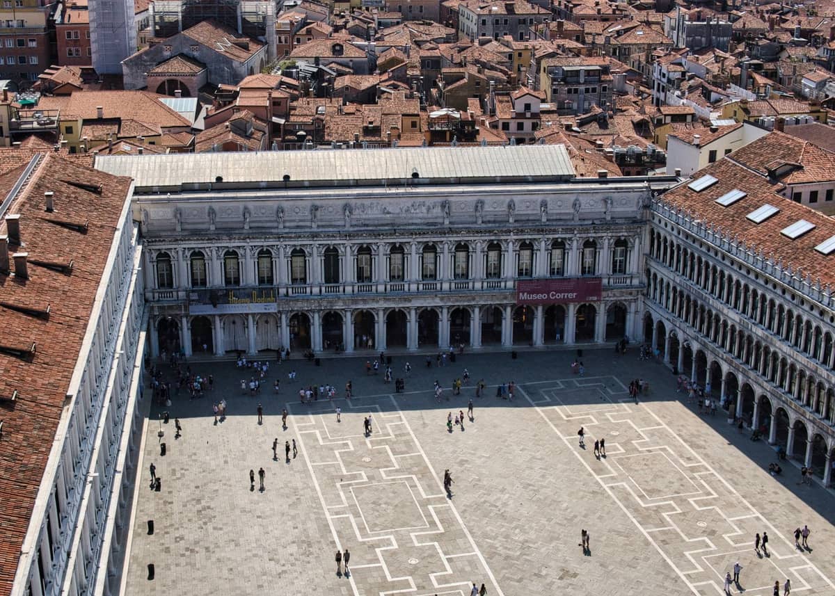 Blick von oben auf die Fassade des Museo Correr über dem Piazza San Marco, mit dem Museumsschild und Passanten auf dem Platz darunter.