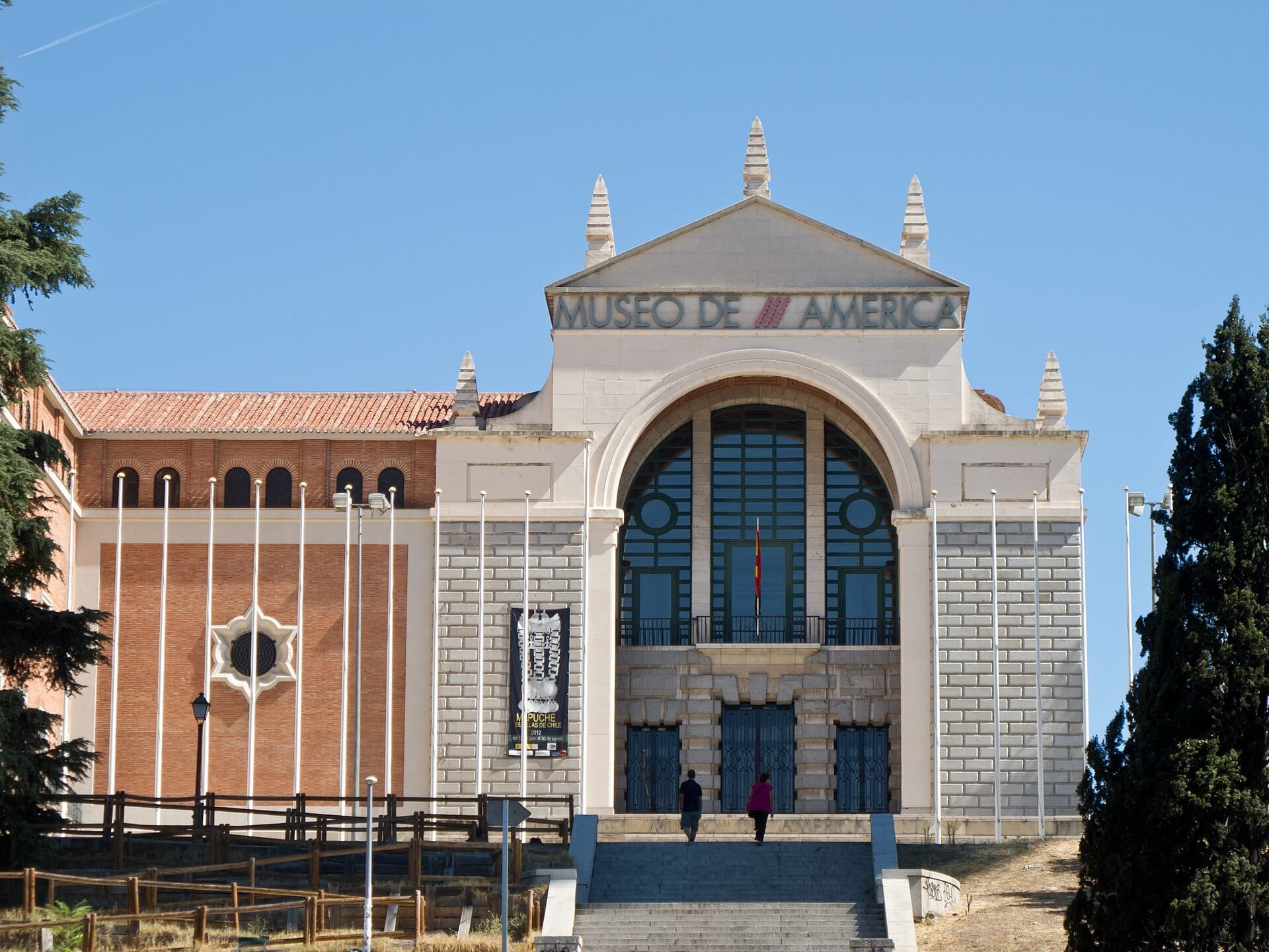 Ingresso principale del Museo de América a Madrid, con una grande facciata in pietra, finestre ad arco e due persone che salgono le scale.
