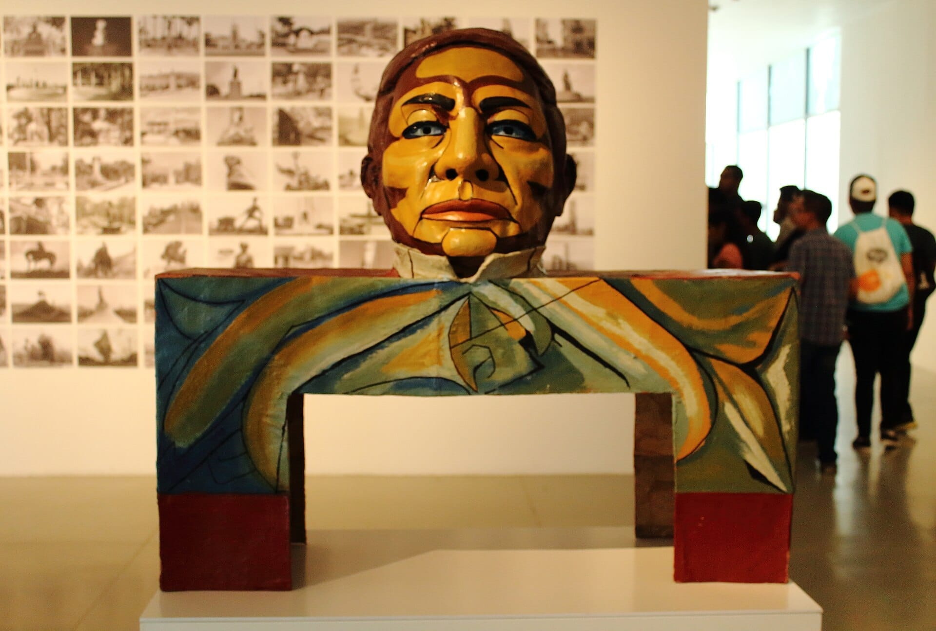 Scultura contemporanea colorata con un viso dipinto esposta al Museo de Arte de Zapopan, con visitatori e una parete di foto sullo sfondo.