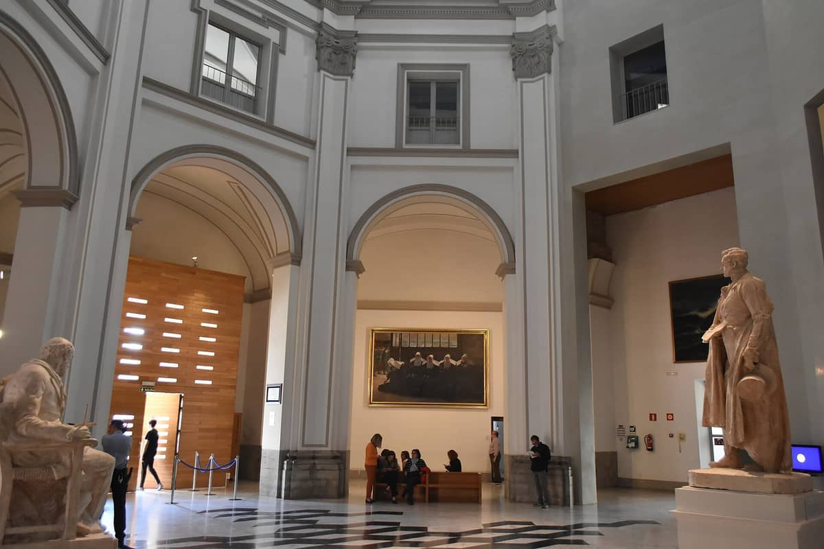 Ampio interno del Museo de Bellas Artes de València, con statue classiche, alte arcate e visitatori che ammirano grandi dipinti.