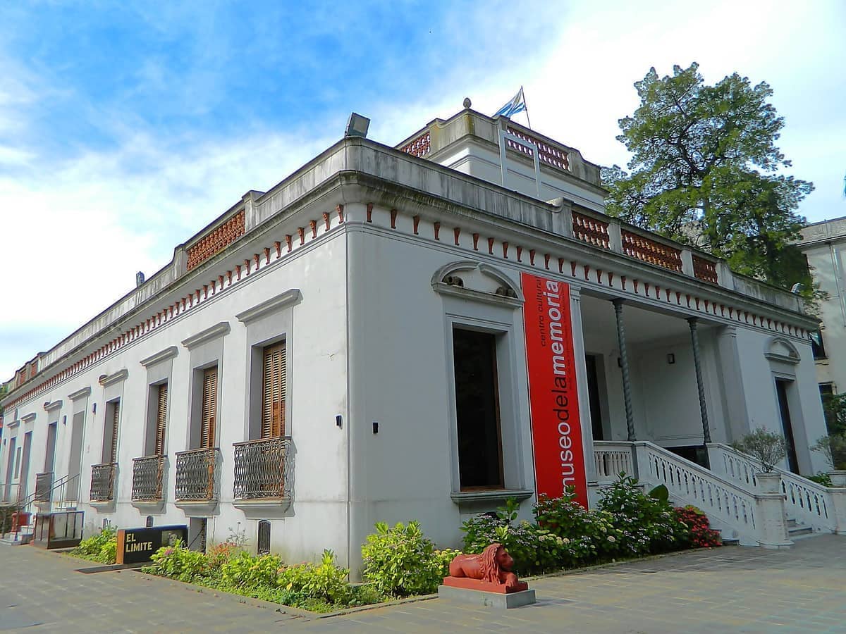 อาคารสีขาวในศตวรรษที่ 19 ของ Museo de la Memoria ในมอนเตวิเดโอ พร้อมป้ายสีแดง รูปปั้นสิงโตหิน และสวนเขียวชอุ่มใต้ท้องฟ้าสีฟ้า