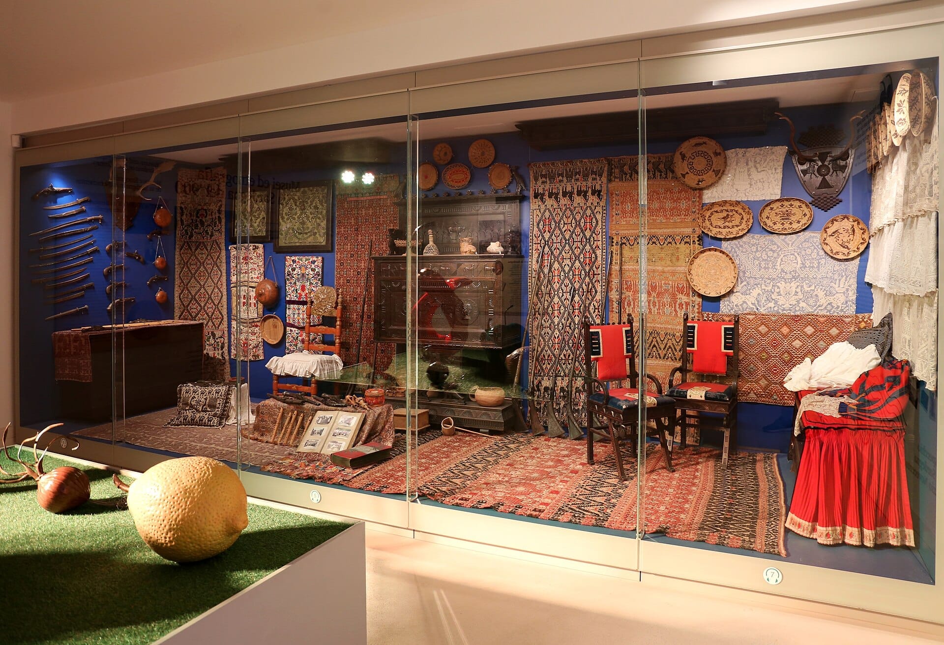 Eine hell erleuchtete Ausstellung im Museo del Costume Nuoro zeigt traditionelle sardische Textilien, Volkstrachten, geschnitzte Holzmöbel und gewebte Körbe hinter Glas.