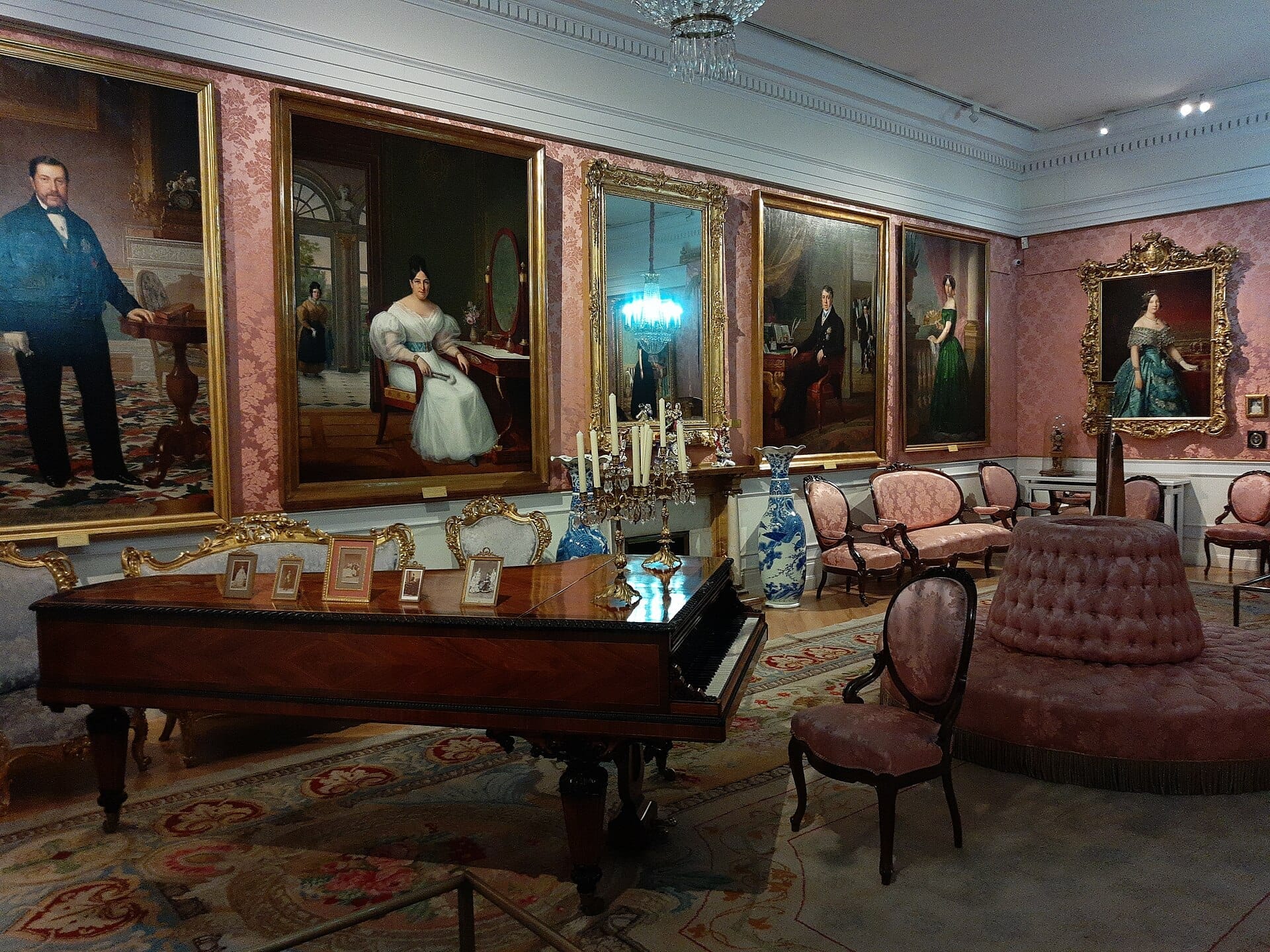 Sontuoso salone del XIX secolo nel Museo del Romanticismo, con mobili antichi, un pianoforte a coda e dipinti dorati con ritratti su pareti tappezzate di rosa.