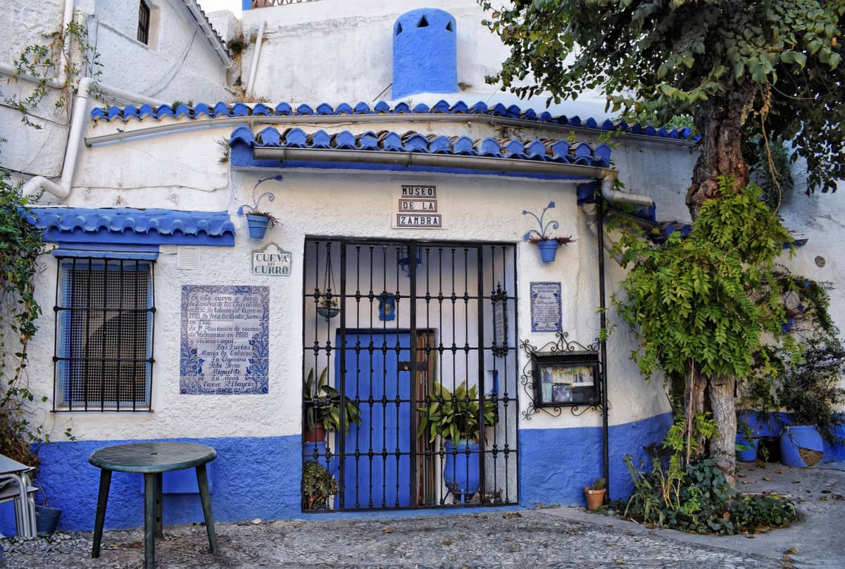 ด้านหน้าพิพิธภัณฑ์ถ้ำ Sacromonte ในกรานาดา บ้านถ้ำปูนขาวแต่งขอบสีน้ำเงิน มีต้นไม้กระถางและกระเบื้องประดับ