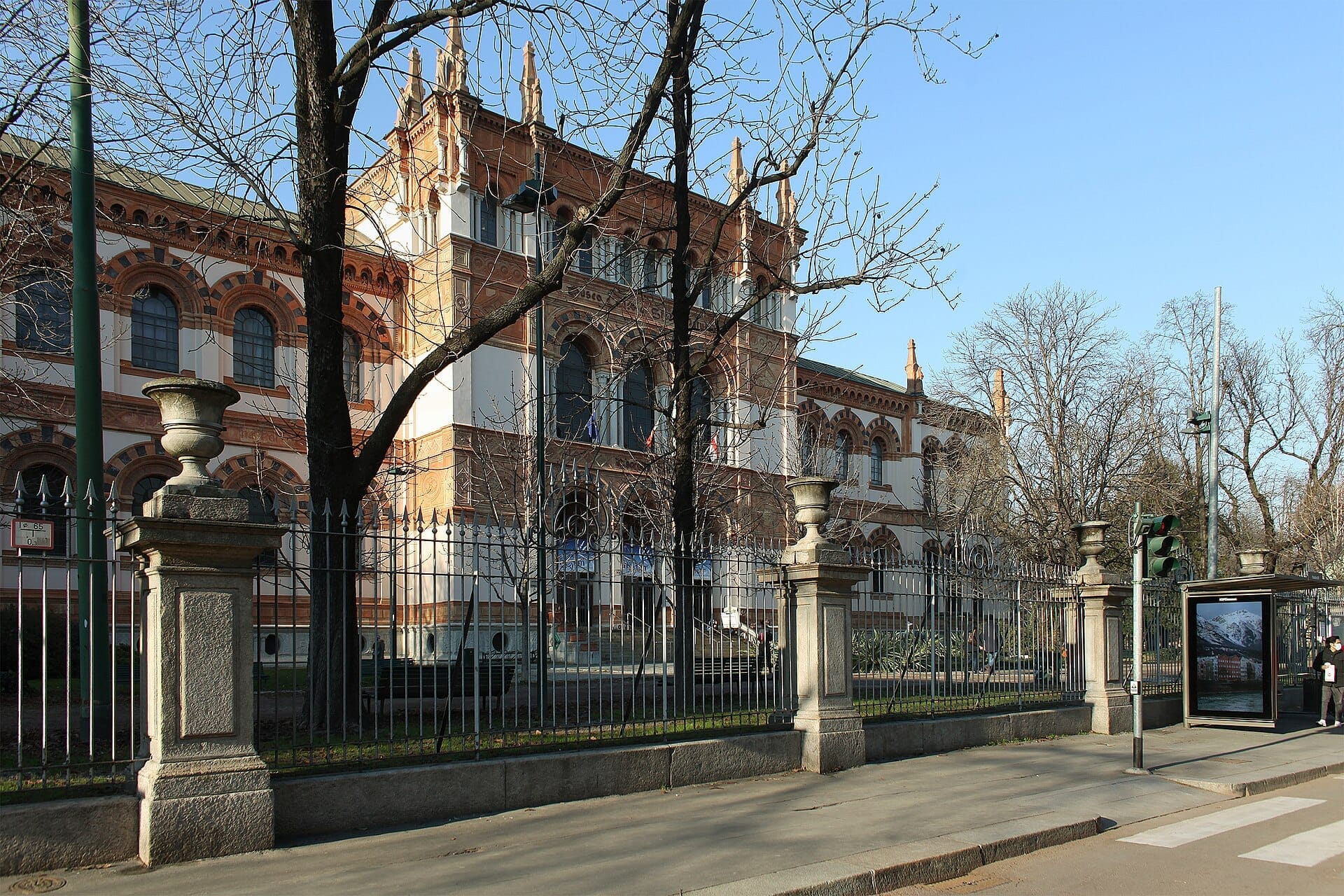 La facciata neogotica del Museo di Storia Naturale di Milano con finestre ad arco, cancellata in ferro, alberi spogli e cielo azzurro limpido.