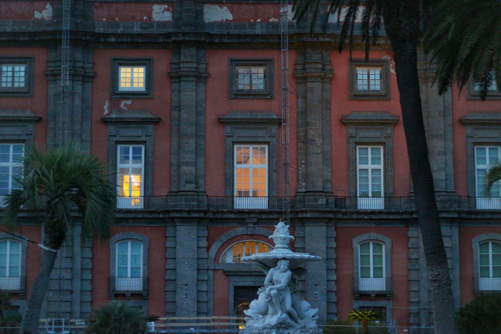 Vista frontale del Museo e Real Bosco di Capodimonte con la sua facciata rossa, le grandi finestre illuminate dall'interno e una fontana classica.