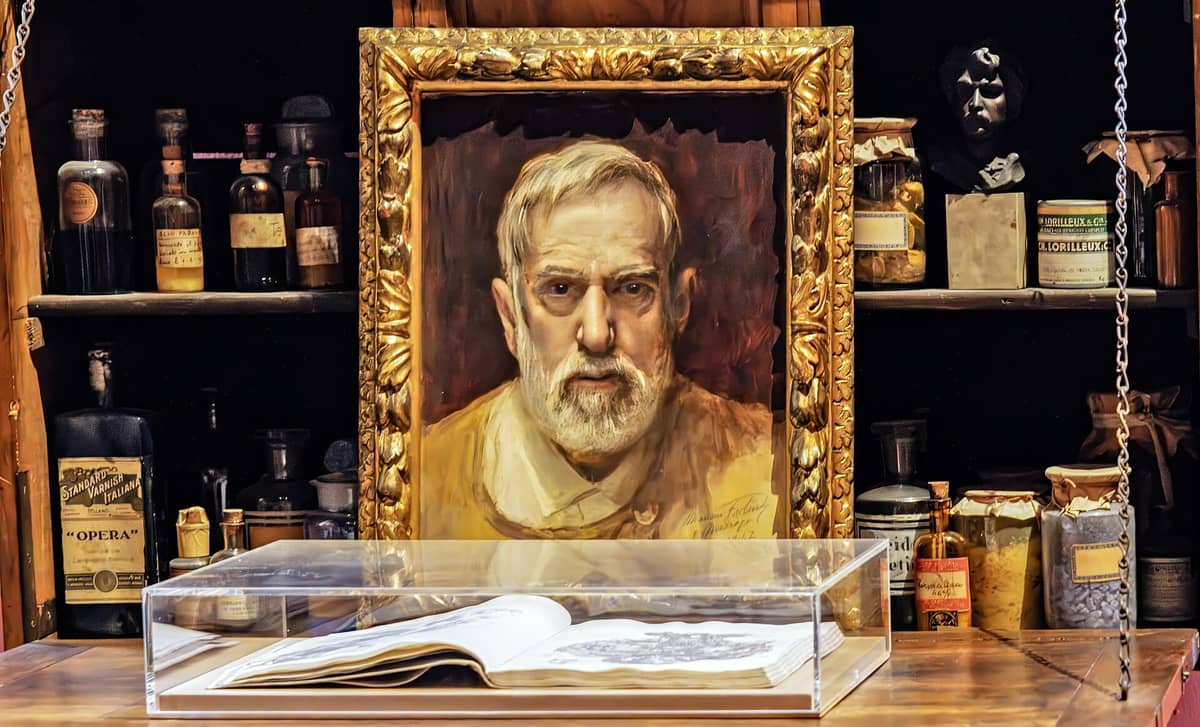 Ein gerahmtes Porträt von Mariano Fortuny im Museo Fortuny, umgeben von Regalen mit alten Flaschen und Objekten – die Atmosphäre eines Künstlerateliers zum Greifen nah.